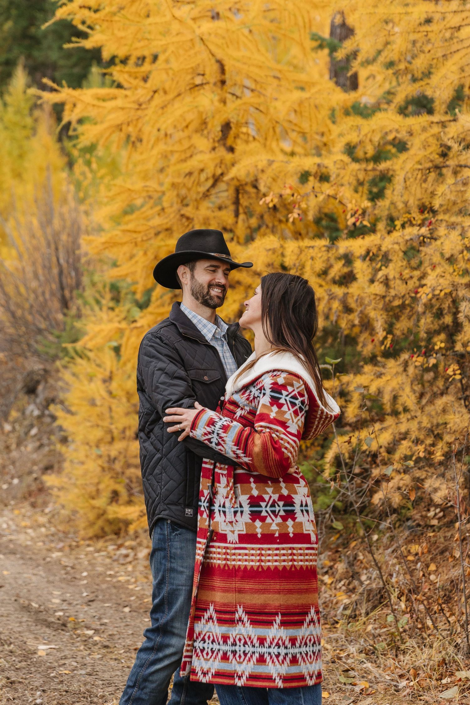 A couple wearing winter sweaters embraces on a path surrounded by golden autumn foliage.