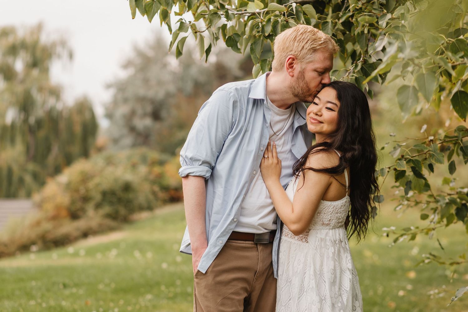 A couple embraces tenderly under green foliage in a sunlit garden setting during a romantic outdoor photoshoot.
