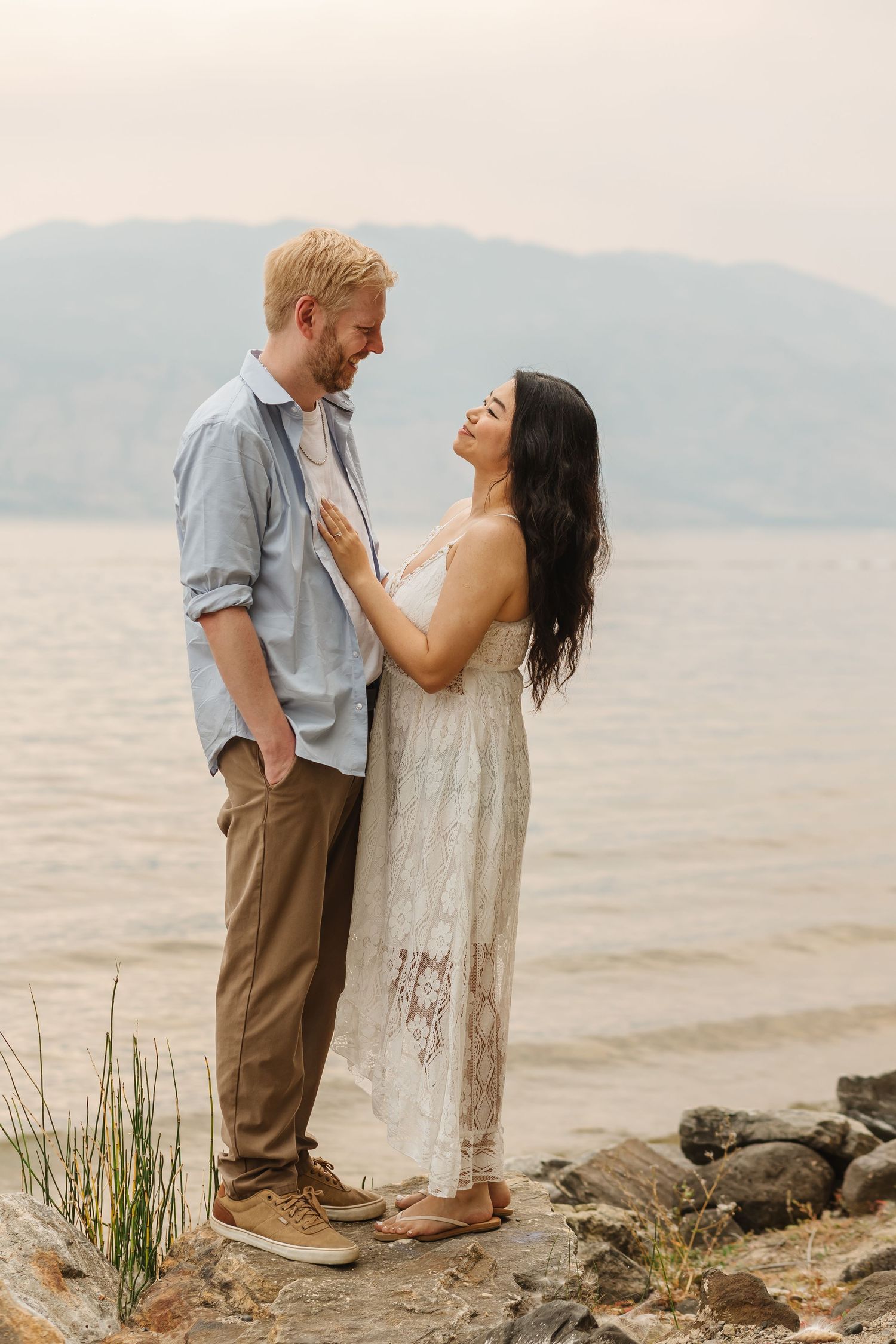 A romantic couple stands together on a rocky beach shoreline with mountains visible in the background.