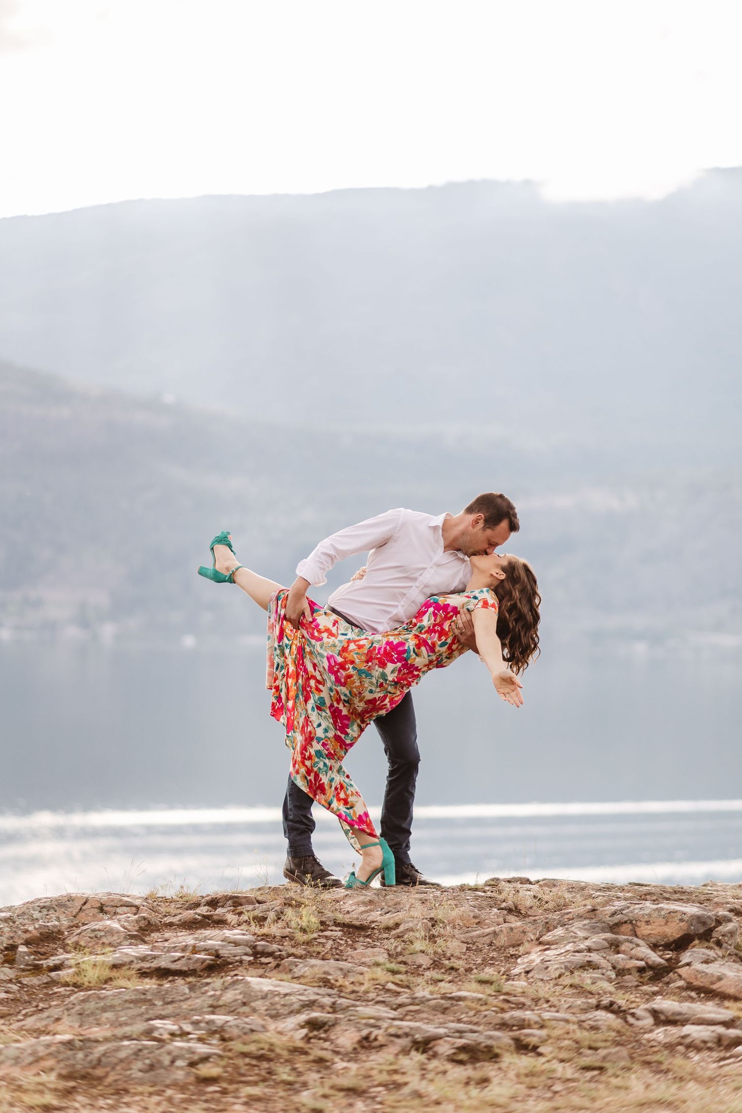 A couple performs playful poses on a cliff overlooking water.