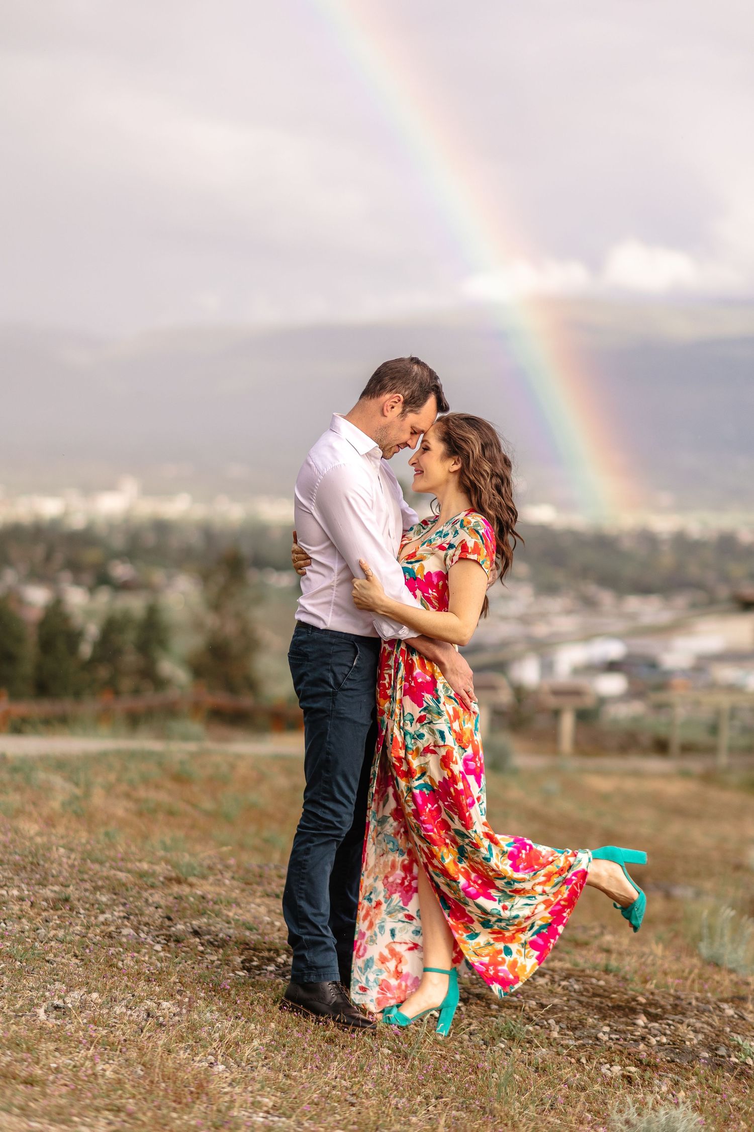 Beautiful rainbow appears behind couple embracing on a grassy hillside.