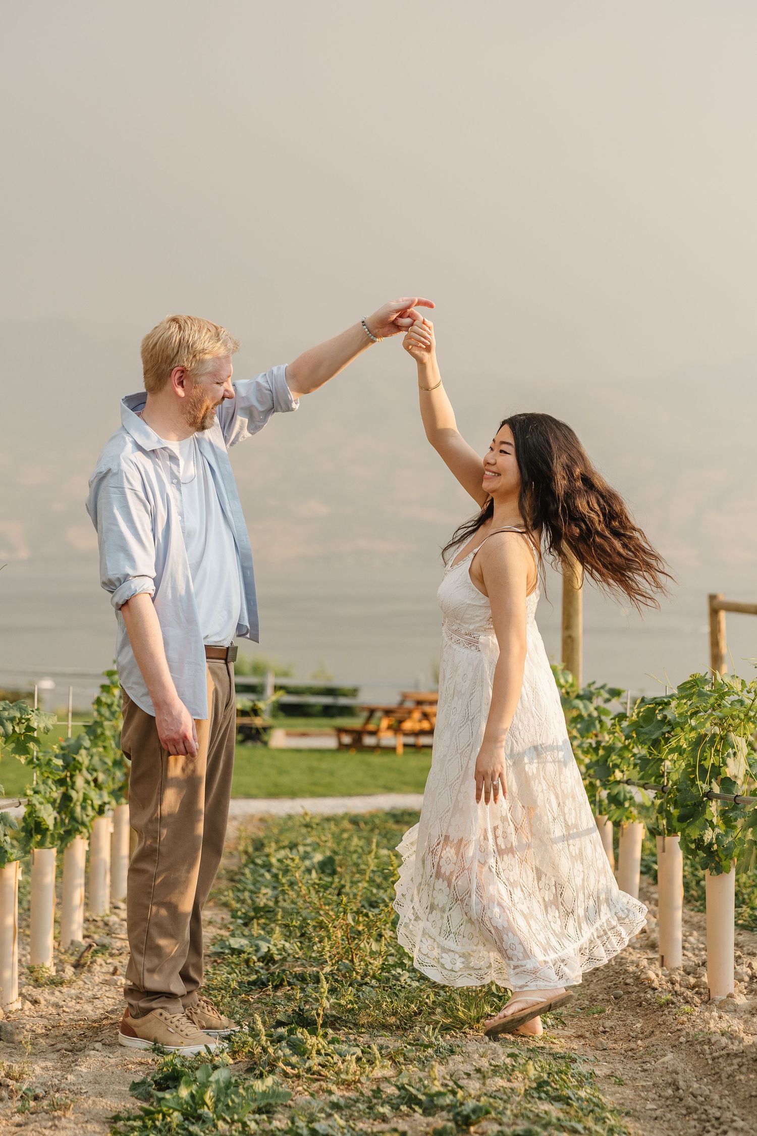 A couple dances together playfully in a vineyard at sunset with white fence posts in the background.