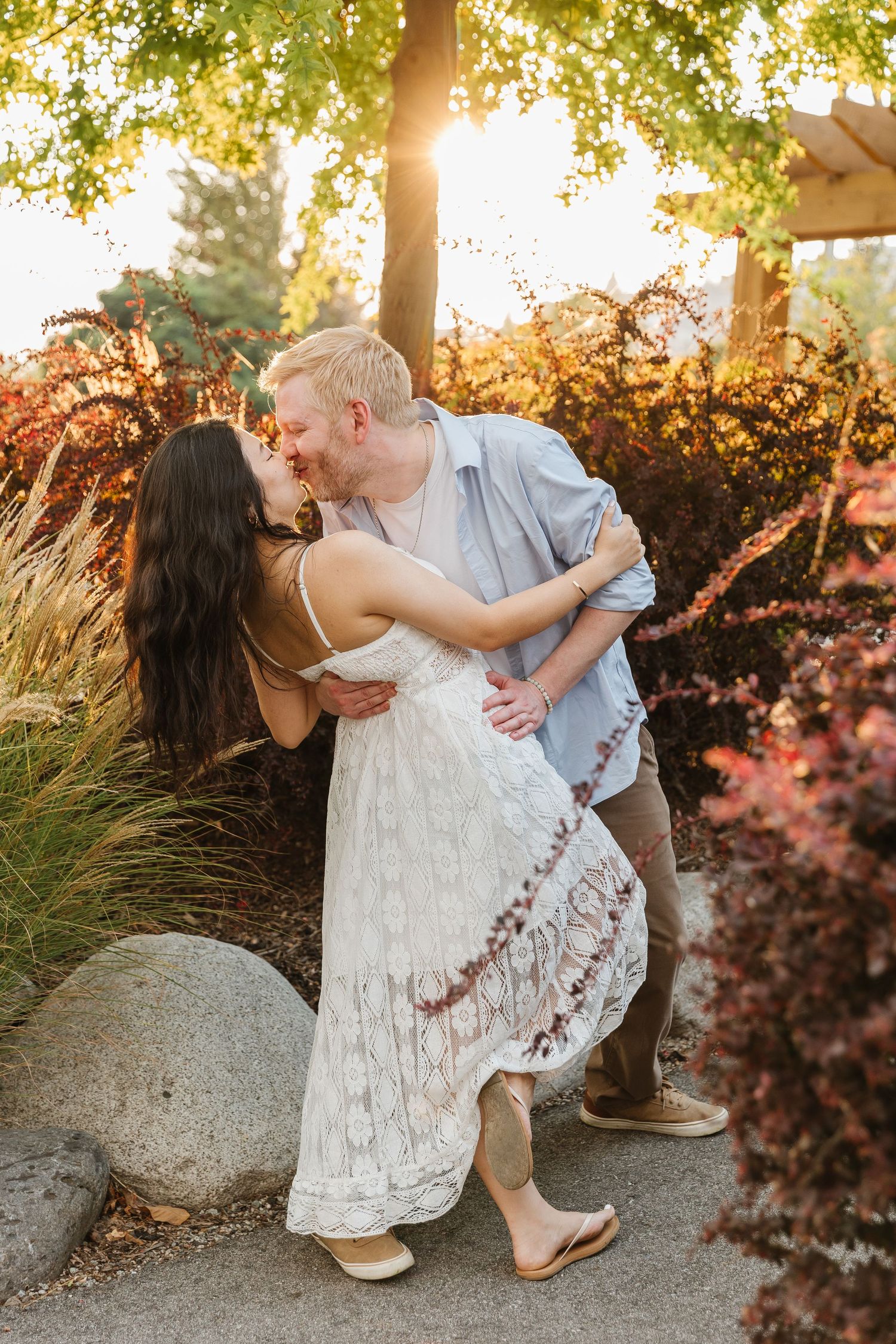 A romantic couple shares a tender dip pose against a sunlit garden backdrop with lush greenery and autumn foliage.