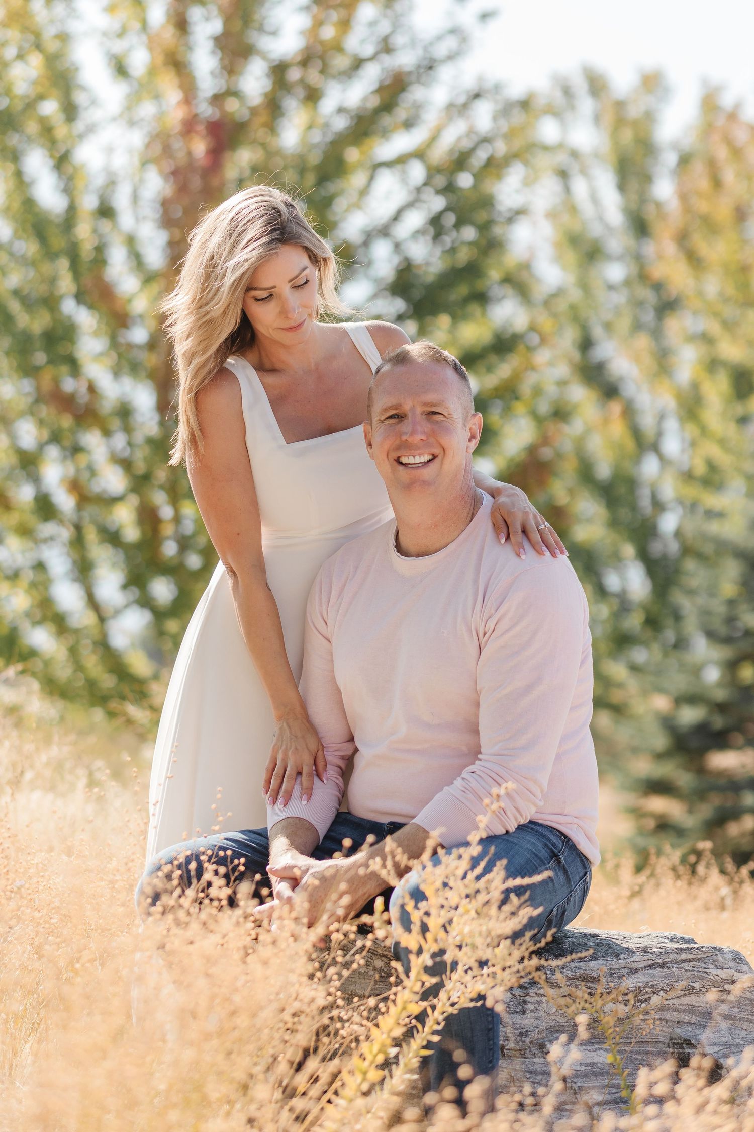 A couple poses together outdoors in a sunlit field with golden grass and evergreen trees in the background.