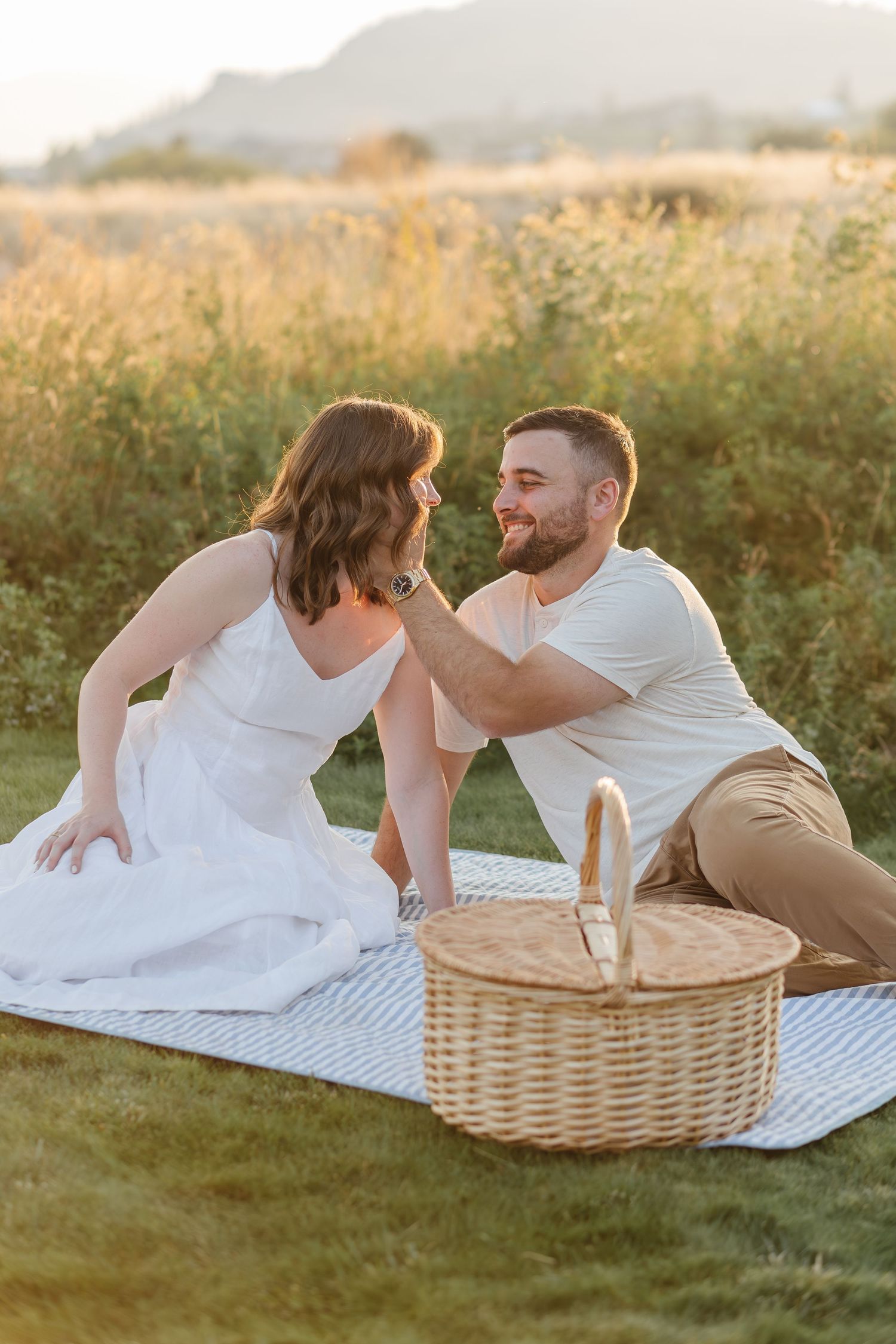 A couple lounges on a white blanket with a picnic basket during golden hour in a grassy field.