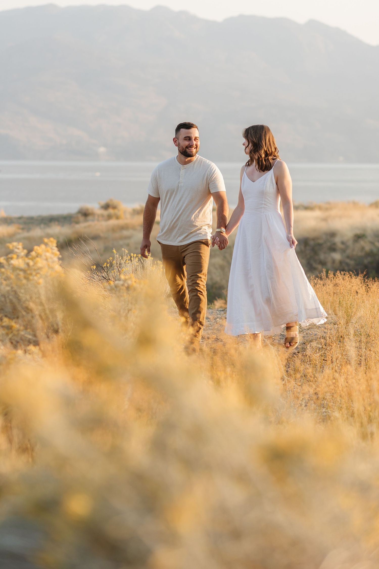 A couple walks hand in hand through golden grass at sunset with mountains in the distance.