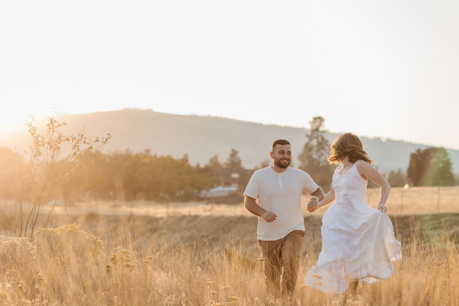A couple in white outfits embraces in a golden sunlit field during sunset with mountains in the background.