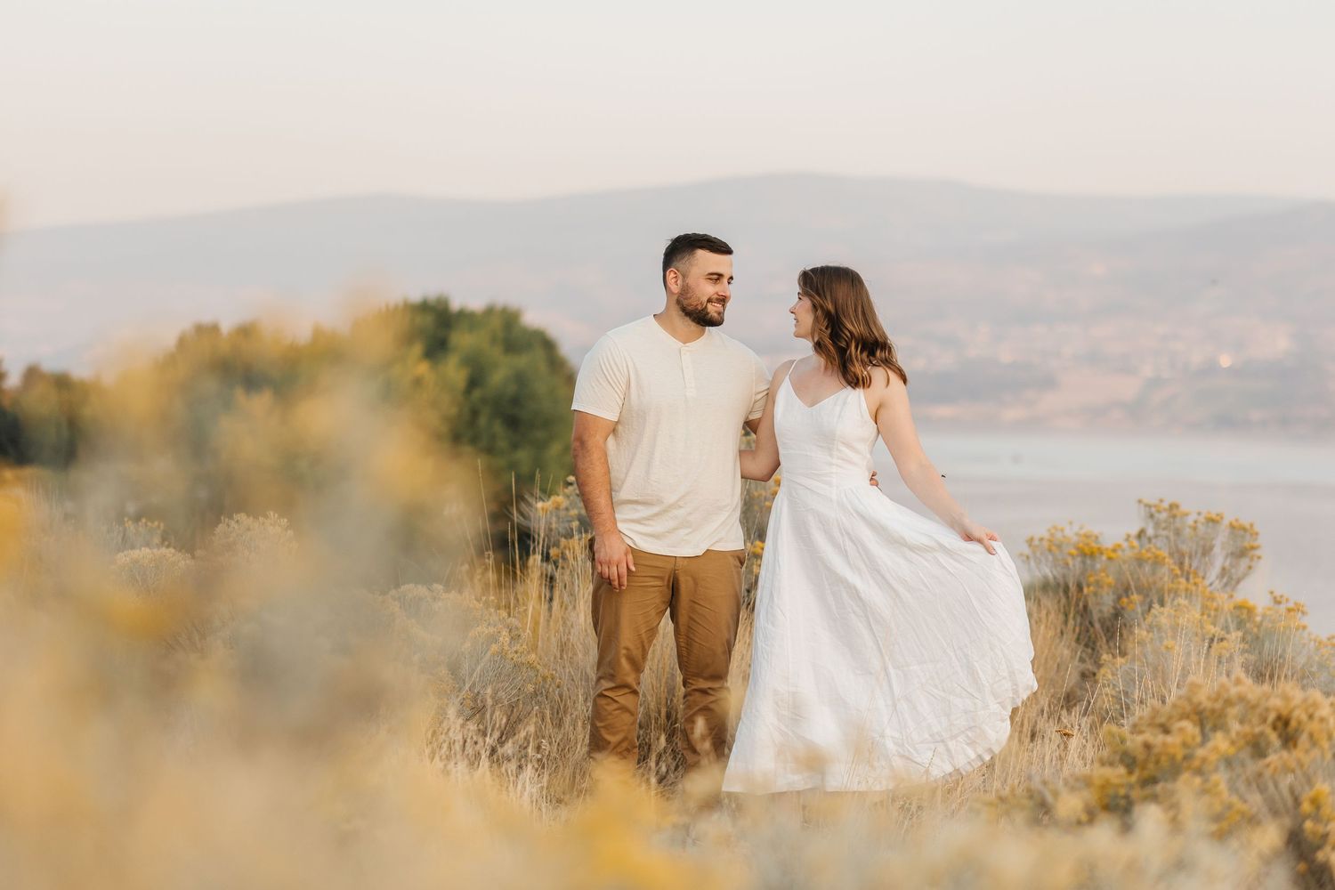 A couple in wedding attire embraces on a hilltop overlooking a scenic mountain landscape during golden hour.