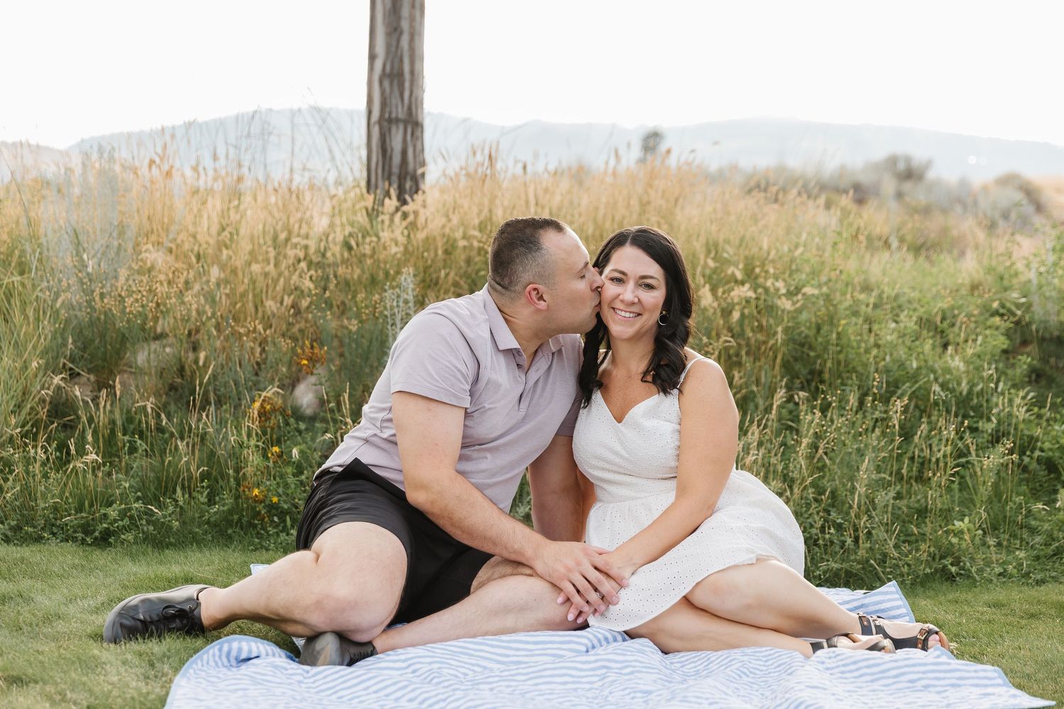 A couple sits together in a grassy meadow during sunset with mountains in the background.