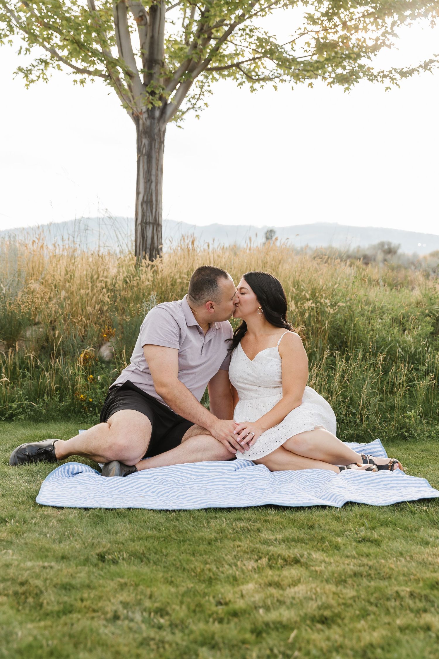 A couple shares a romantic moment while lying on a blanket in a grassy field during golden hour.