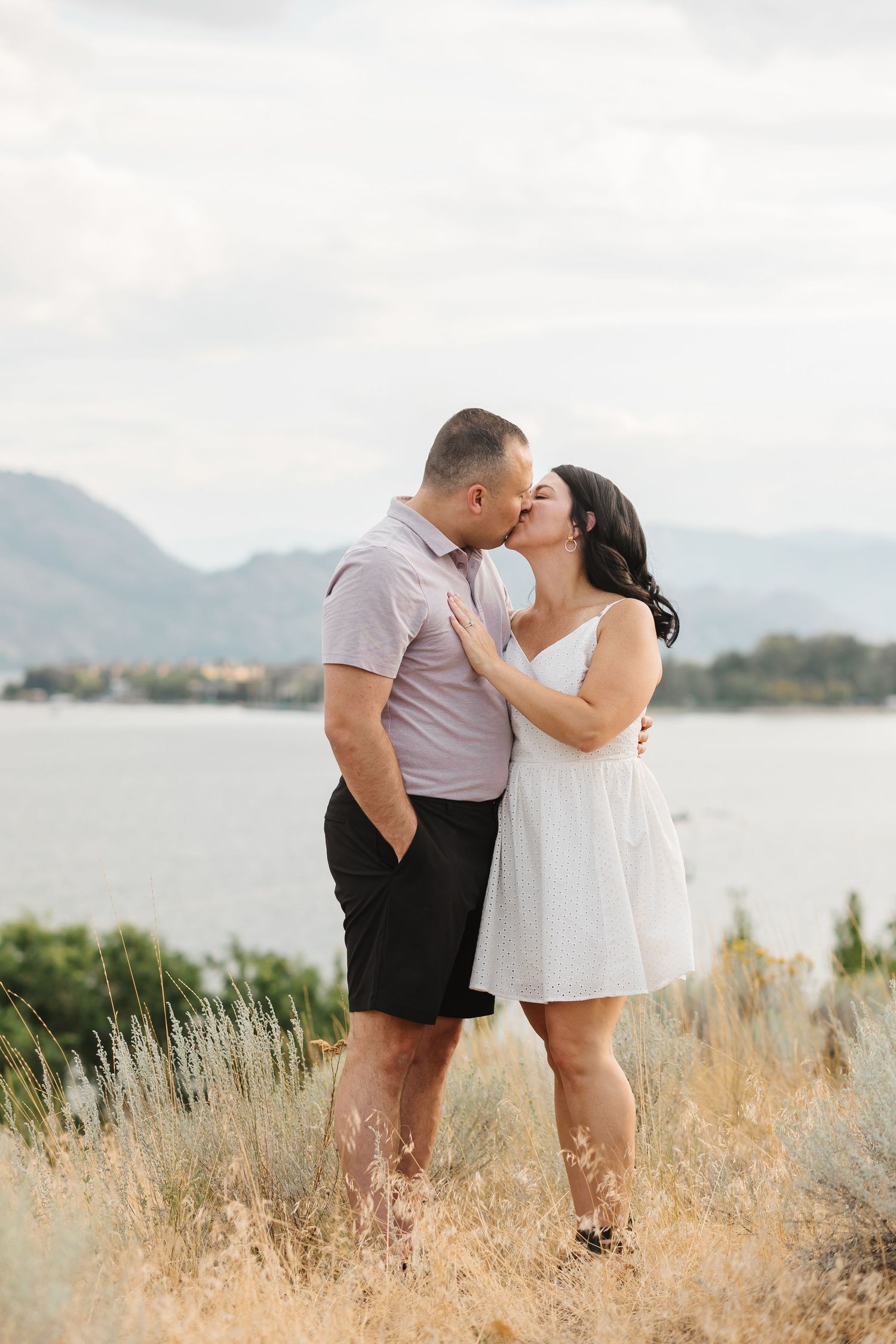 A romantic couple shares a tender kiss in a field of tall grass with a serene lake and mountains in the background.