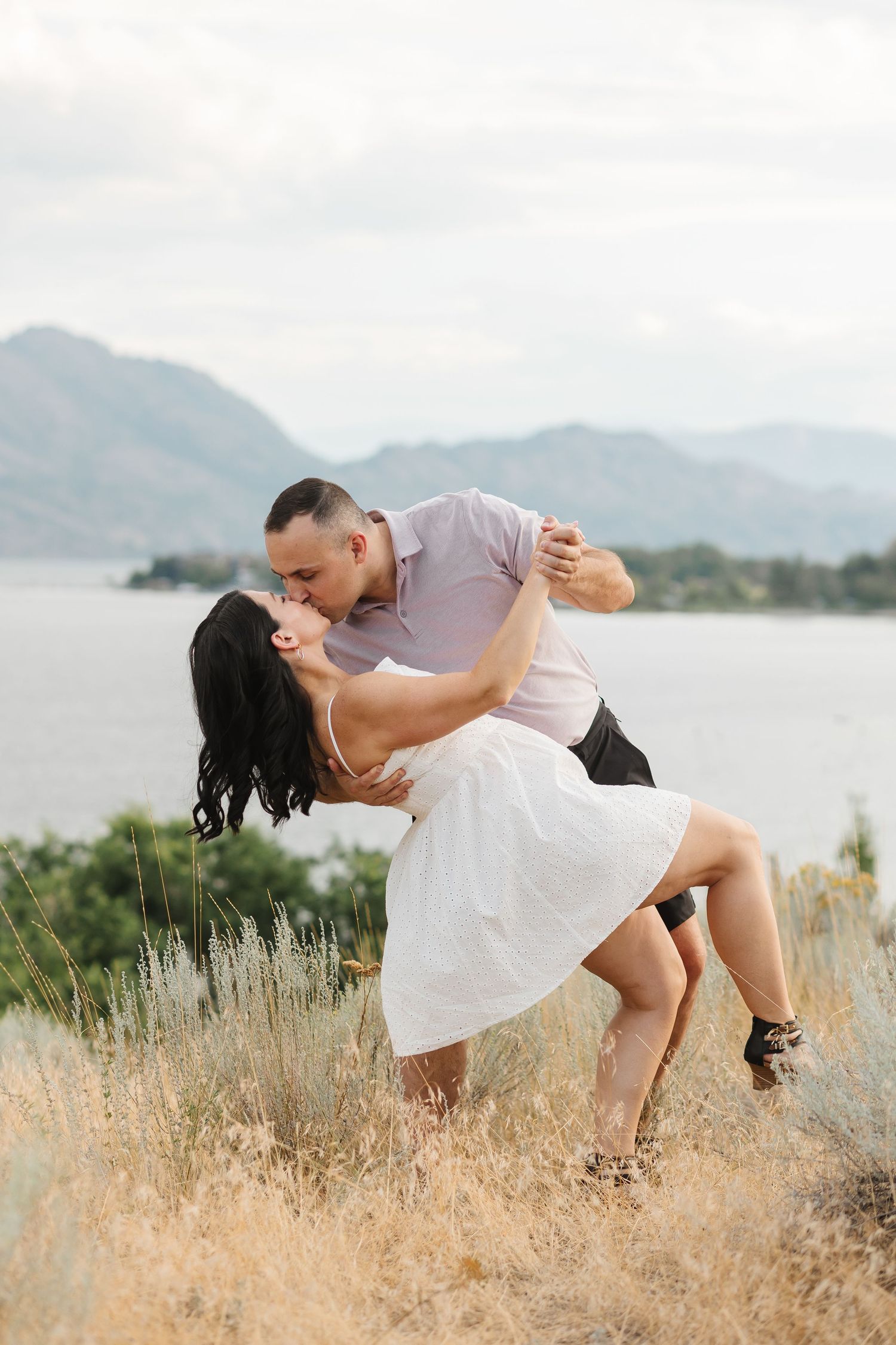 A romantic couple in white outfits dips and kisses against a scenic mountain lake backdrop during a photoshoot.