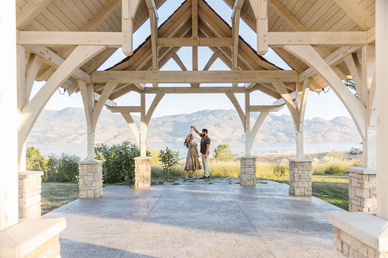 A scenic mountain vista viewed through an elegant wooden pavilion with stone pillars at sunset.