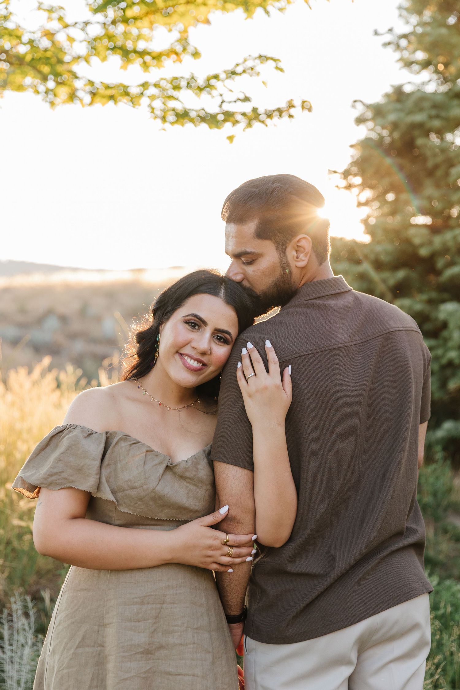 A romantic couple embraces in a golden sunset, with the woman wearing a beige dress and the man in casual attire.
