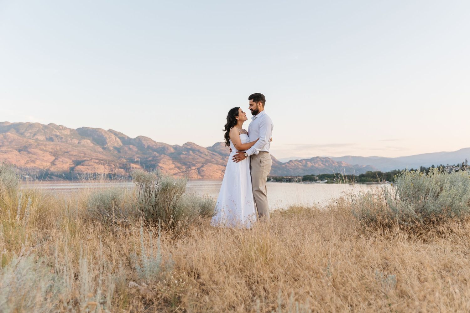 A couple in wedding attire embrace in a golden field with mountains in the background during sunset.