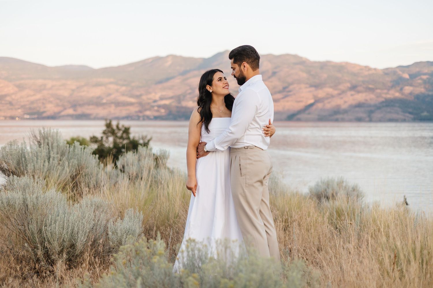 A couple in white embraces against a scenic mountain lake backdrop with desert brush in the foreground.