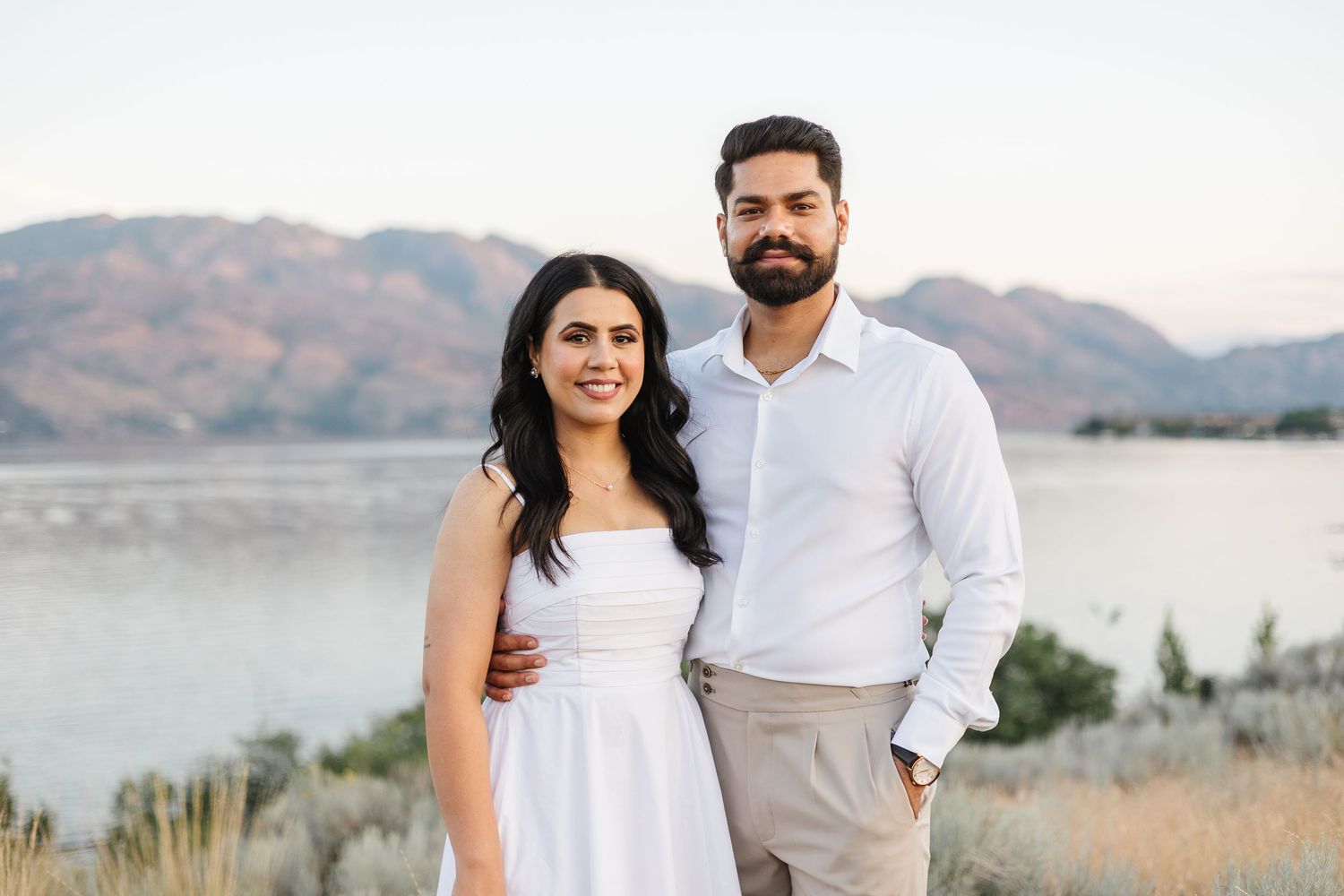 A couple poses together in white outfits against a scenic mountain lake backdrop at sunset.