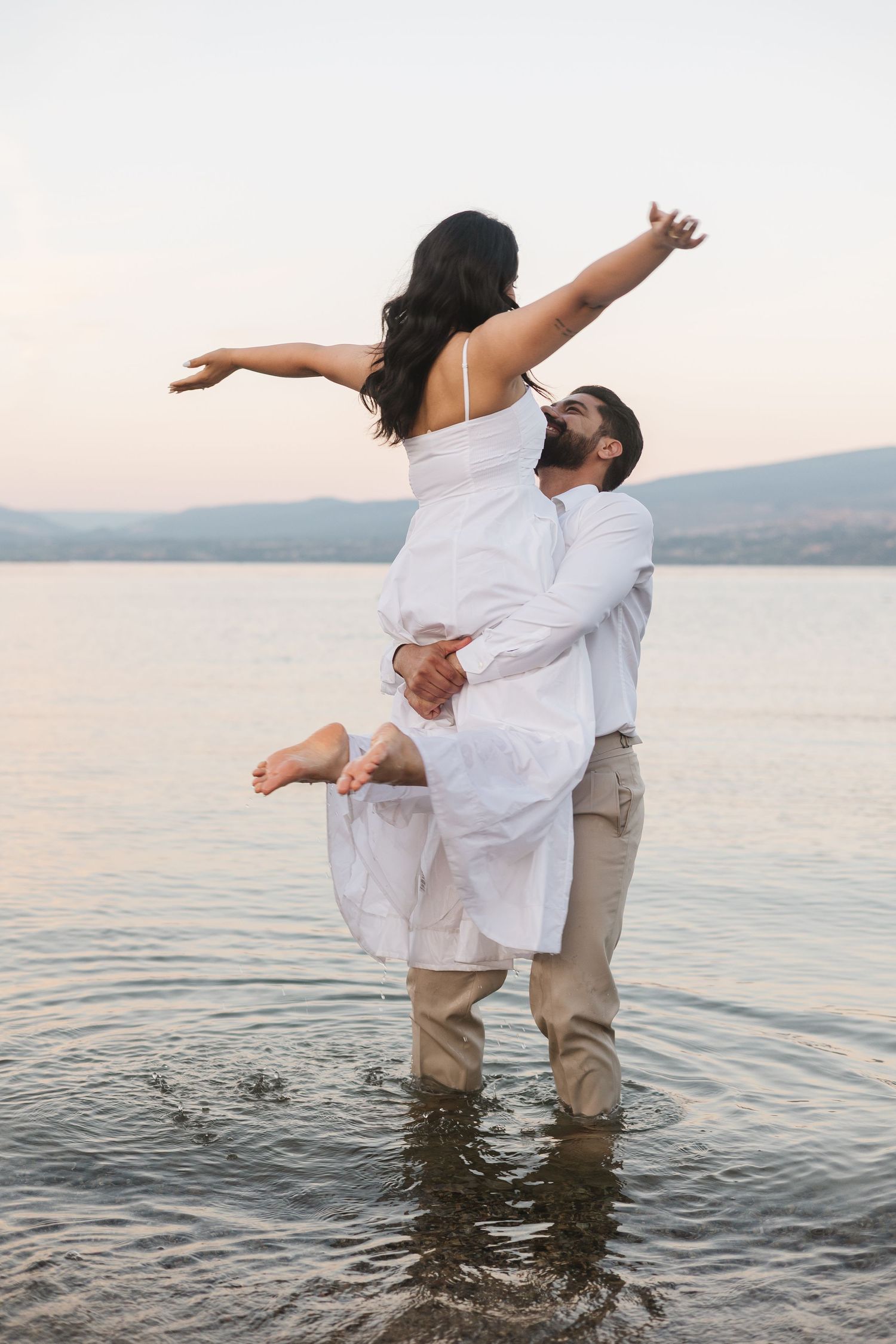 A couple in white and beige clothing stands in shallow water at sunset, creating a romantic silhouette against the lake.