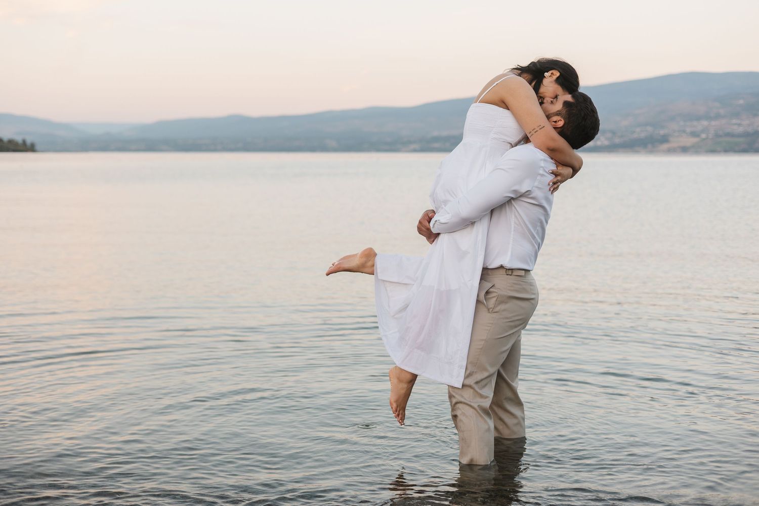 A couple embraces on a beach at sunset with calm waters and mountains in the background.