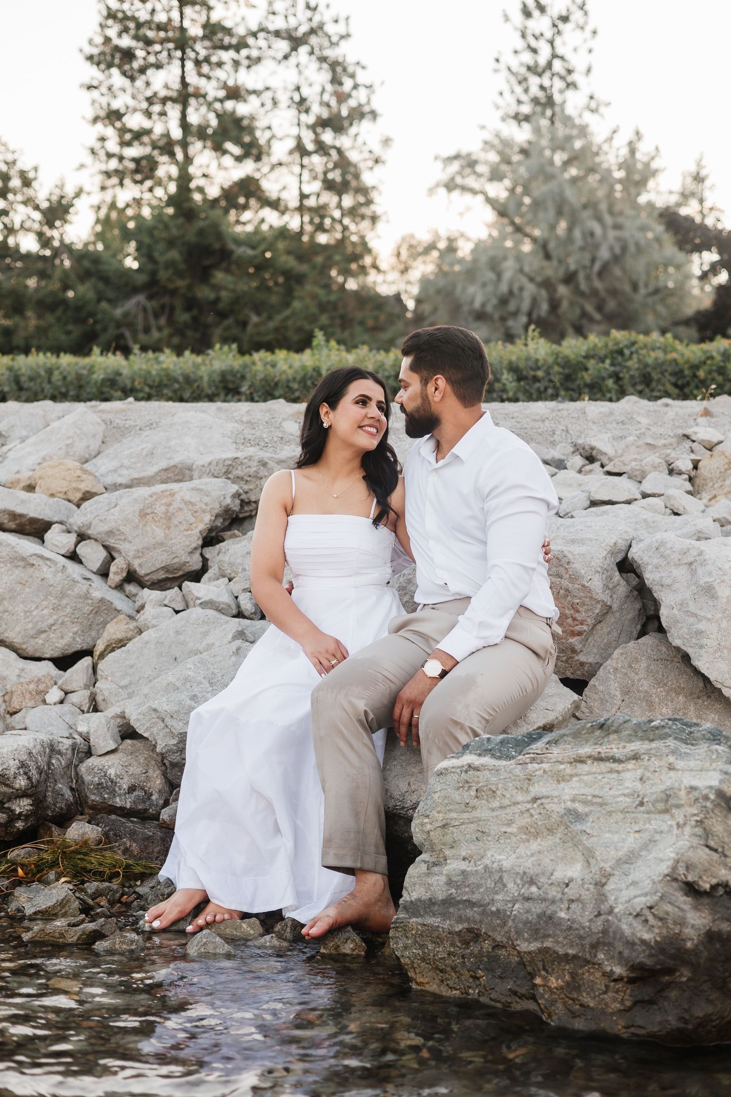 A couple in white and beige outfits sits romantically together on coastal rocks by the water at sunset.