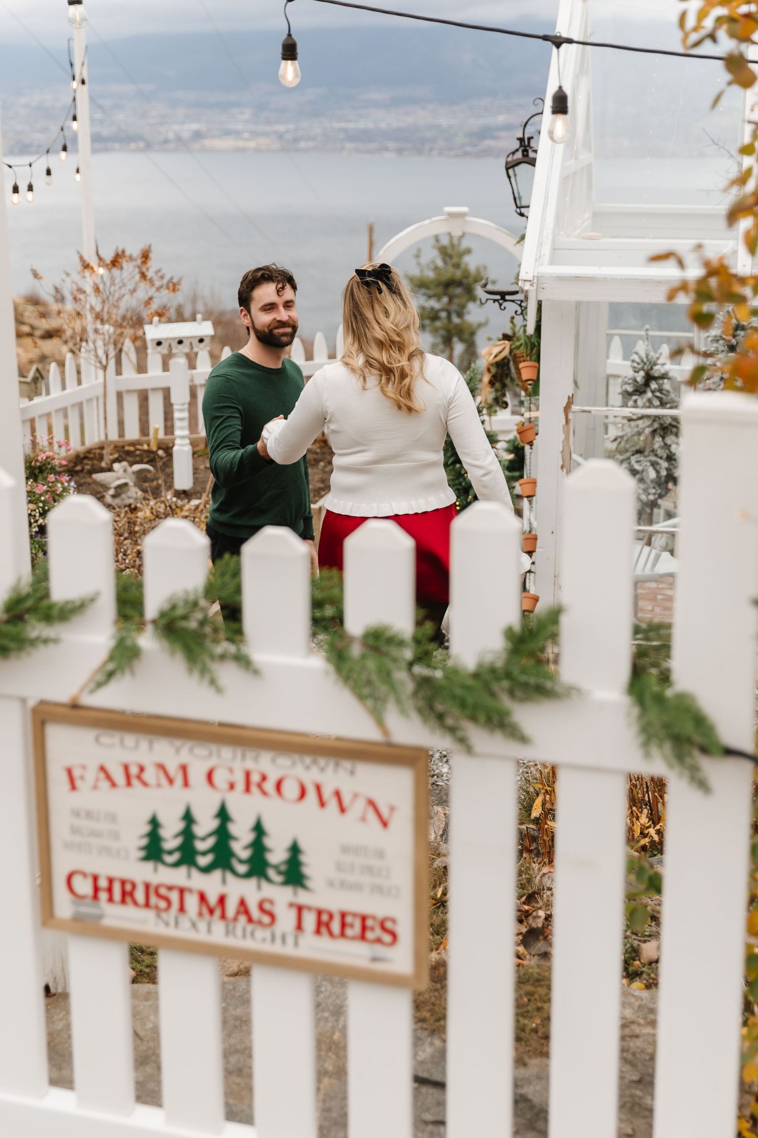 Rustic Christmas tree farm sign hangs on white picket fence with evergreen garland decorations.