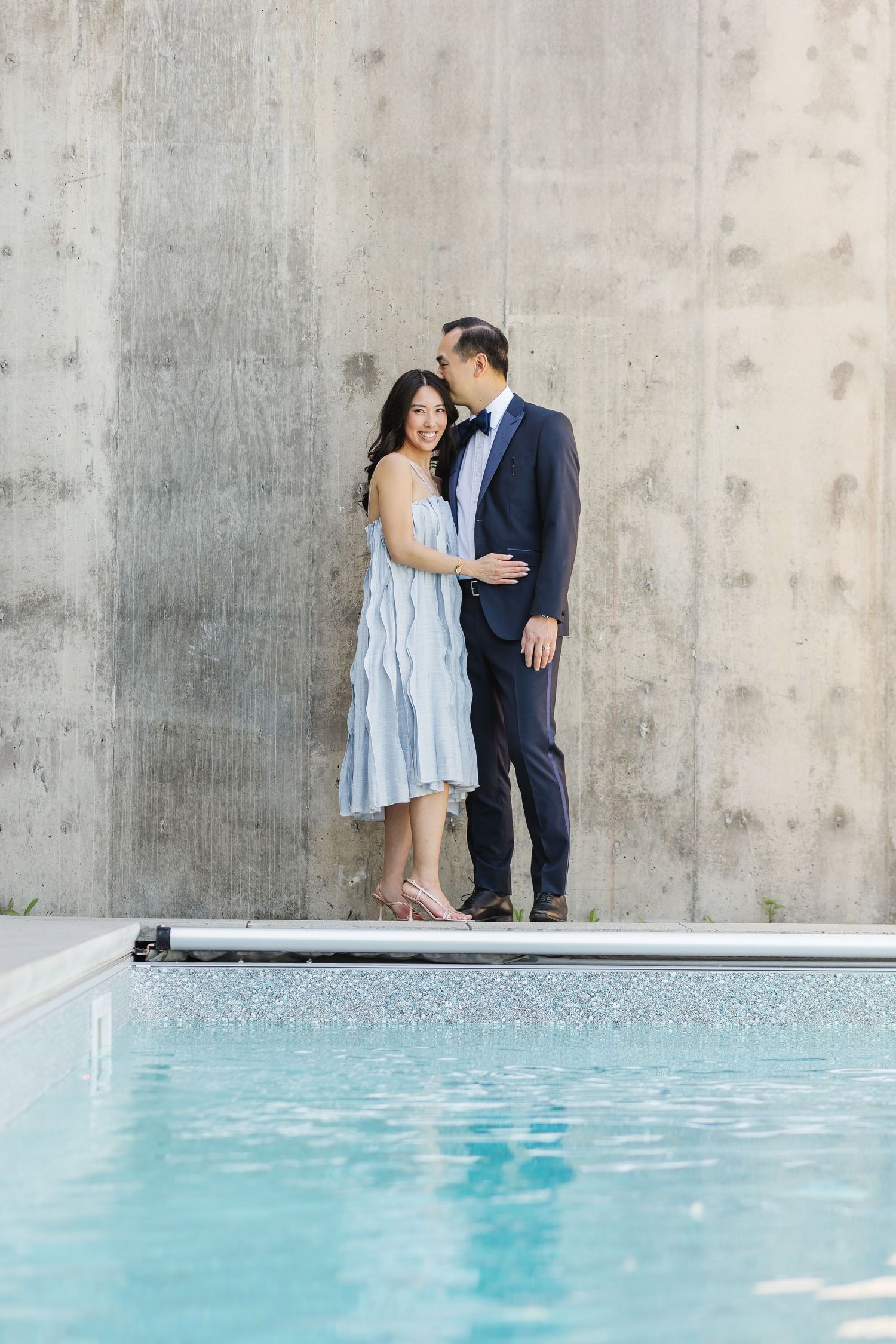 A romantic couple shares an intimate moment against a concrete wall beside a serene turquoise pool.