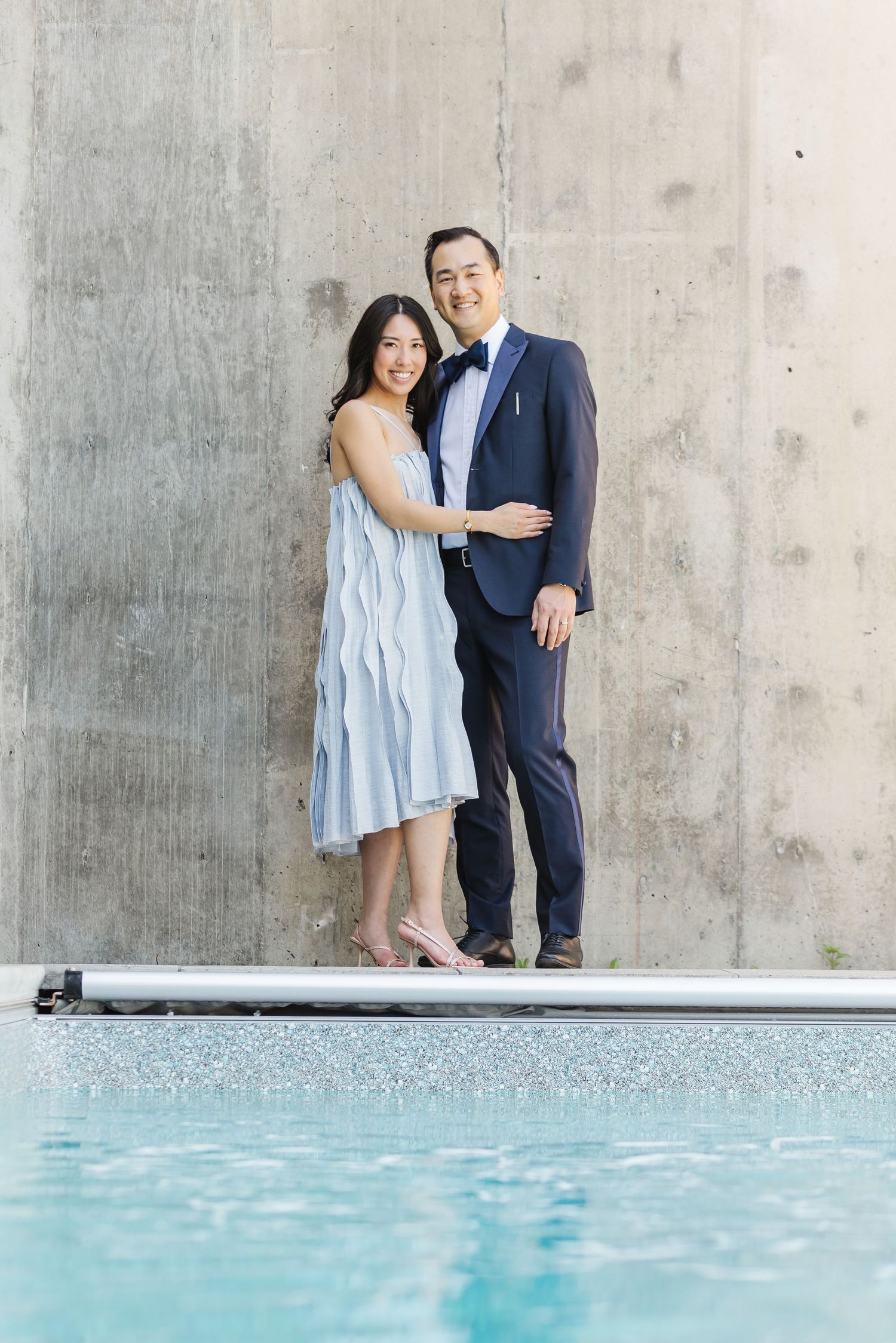 A couple in formal attire poses intimately against a concrete wall near a tranquil swimming pool.