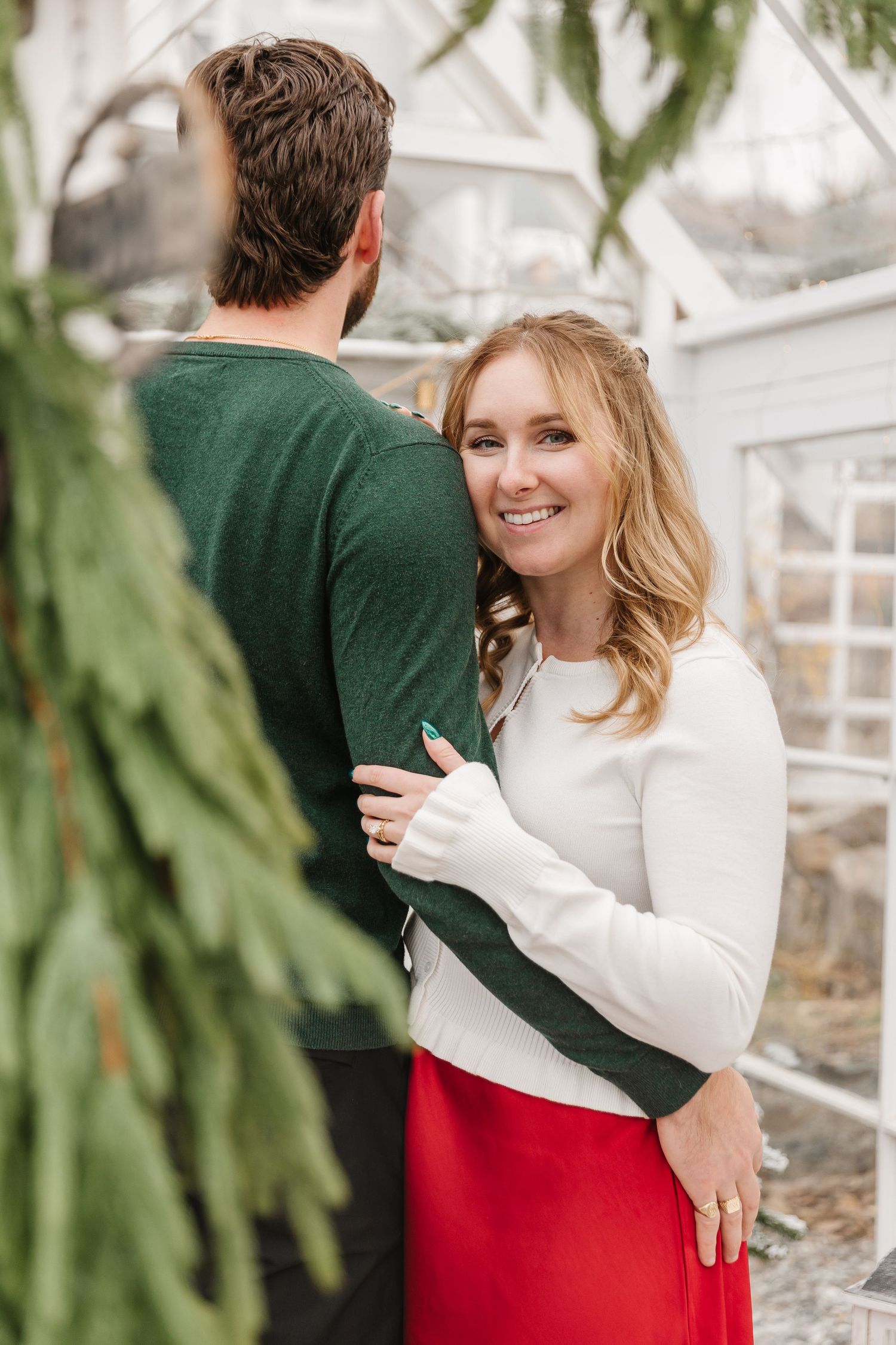 A couple in festive winter attire embraces in a greenhouse setting with greenery visible in the background.
