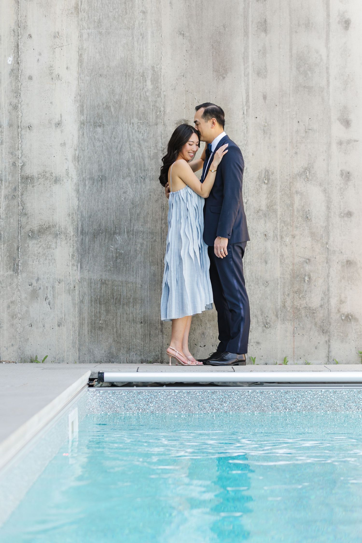 A romantic couple shares intimate moments by a swimming pool against a concrete wall backdrop.