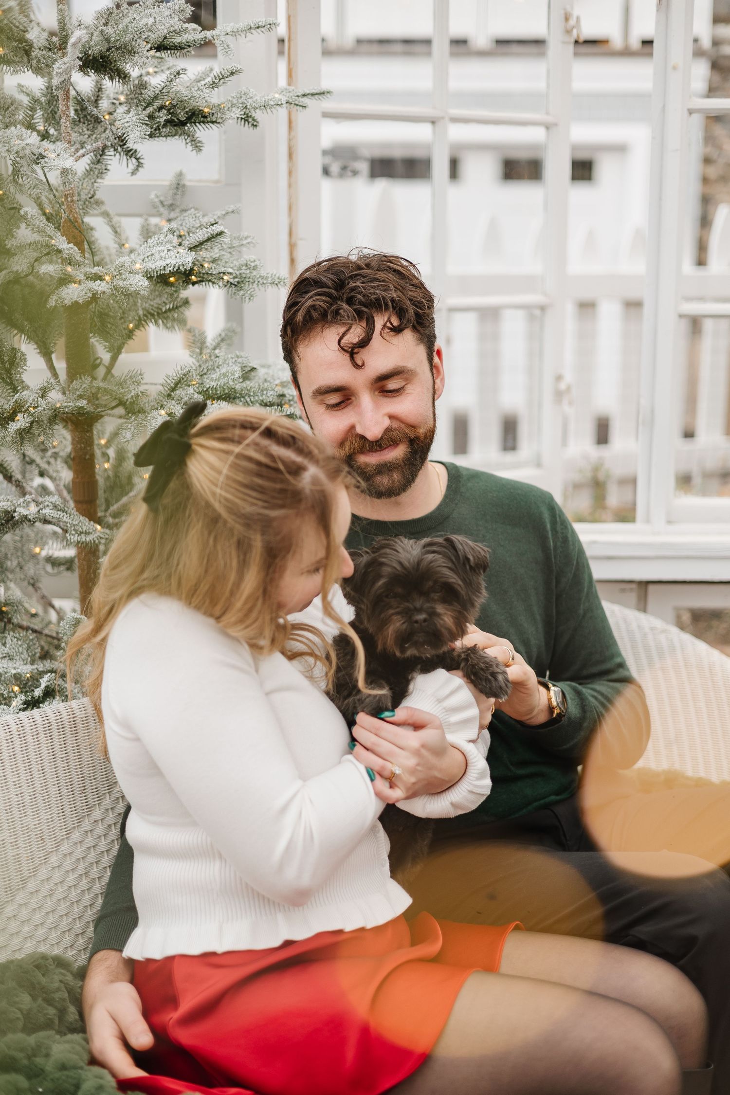 Couple sitting on couch with small black dog during holidays with Christmas tree in background.