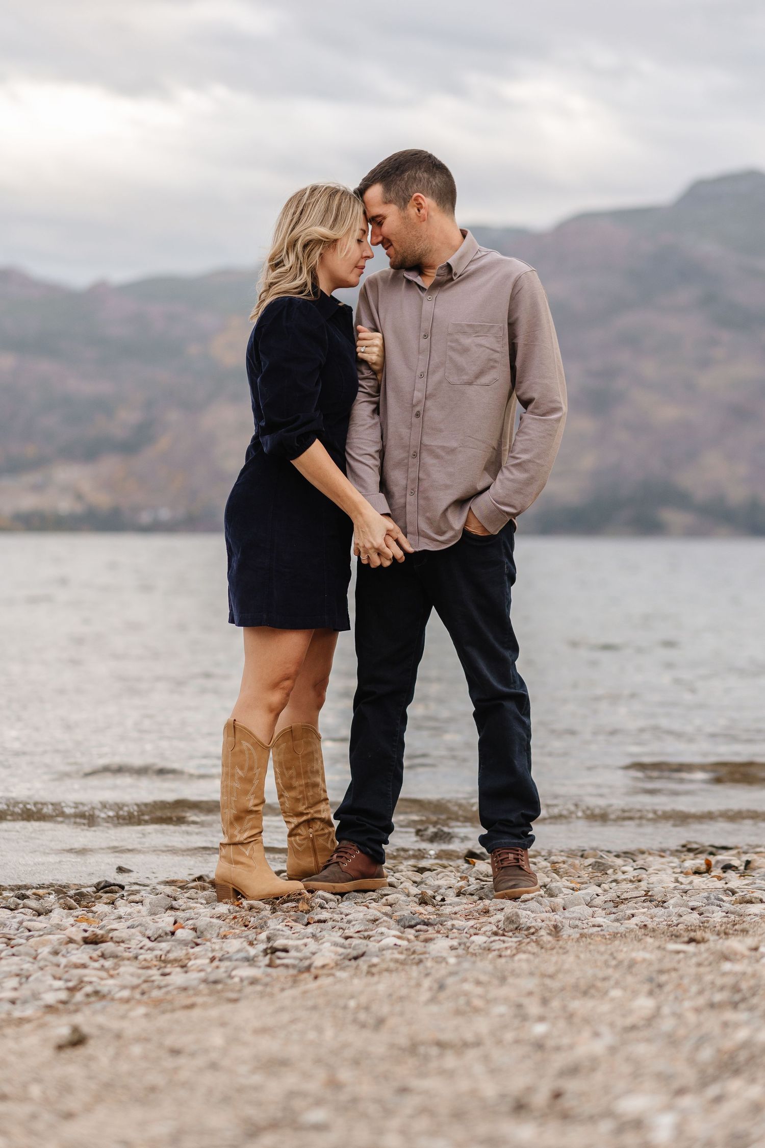 Couple sharing intimate moment on rocky beach with mountains and lake in background.