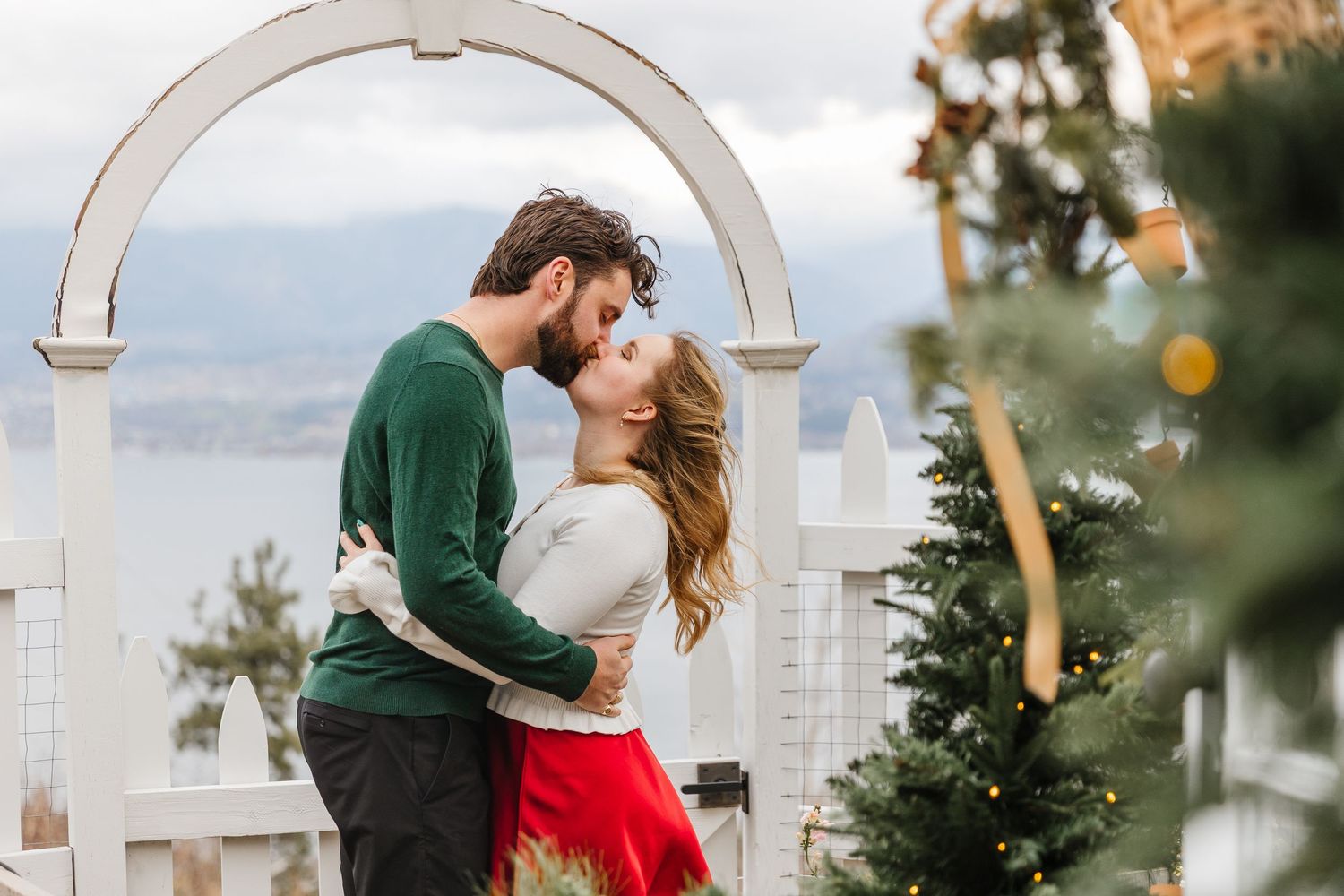 Couple in romantic embrace shares tender moment against waterfront backdrop with Christmas trees and twinkling lights.