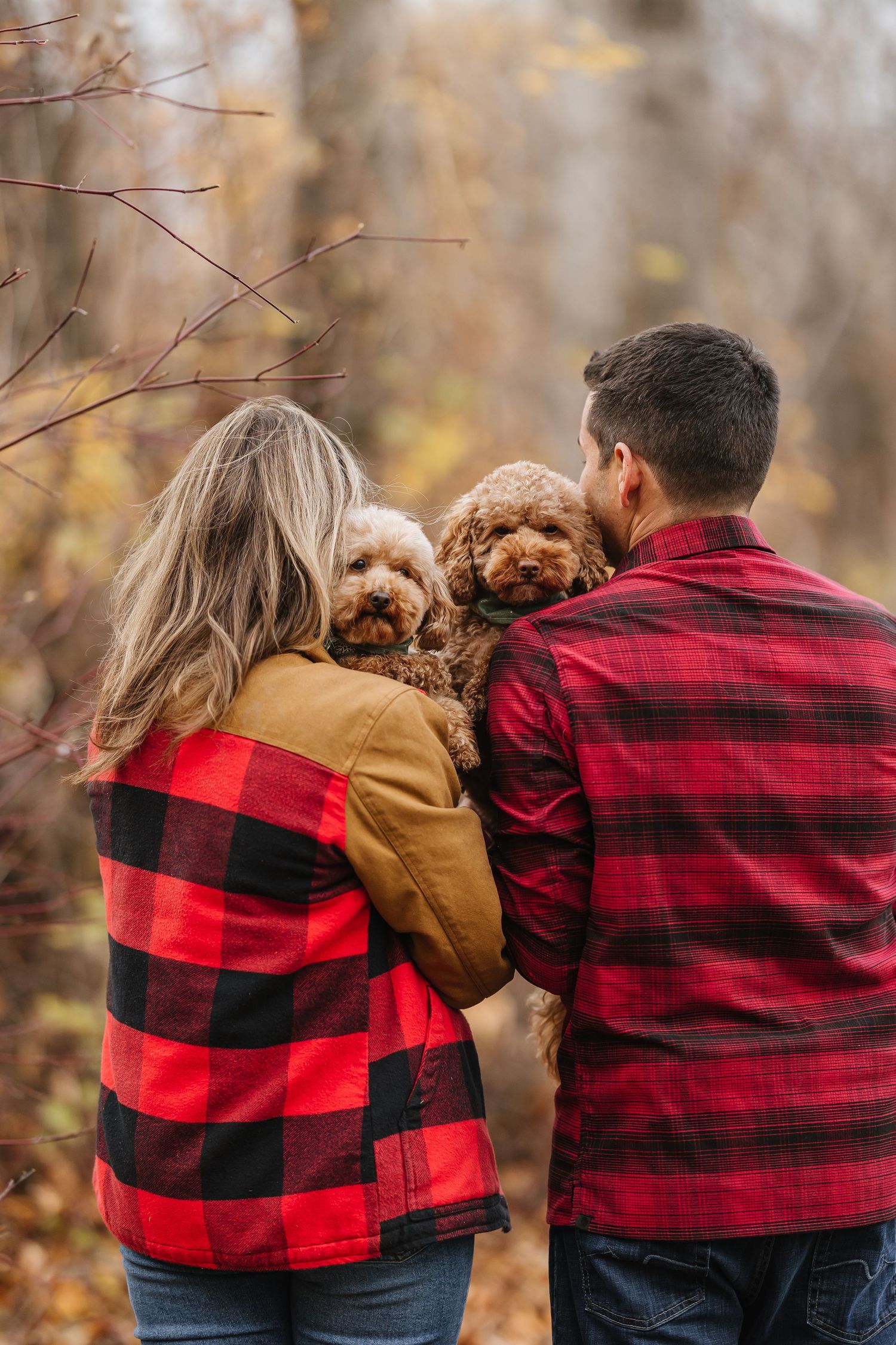 A couple in matching buffalo plaid jackets walk together with their small brown dog in an autumn setting.