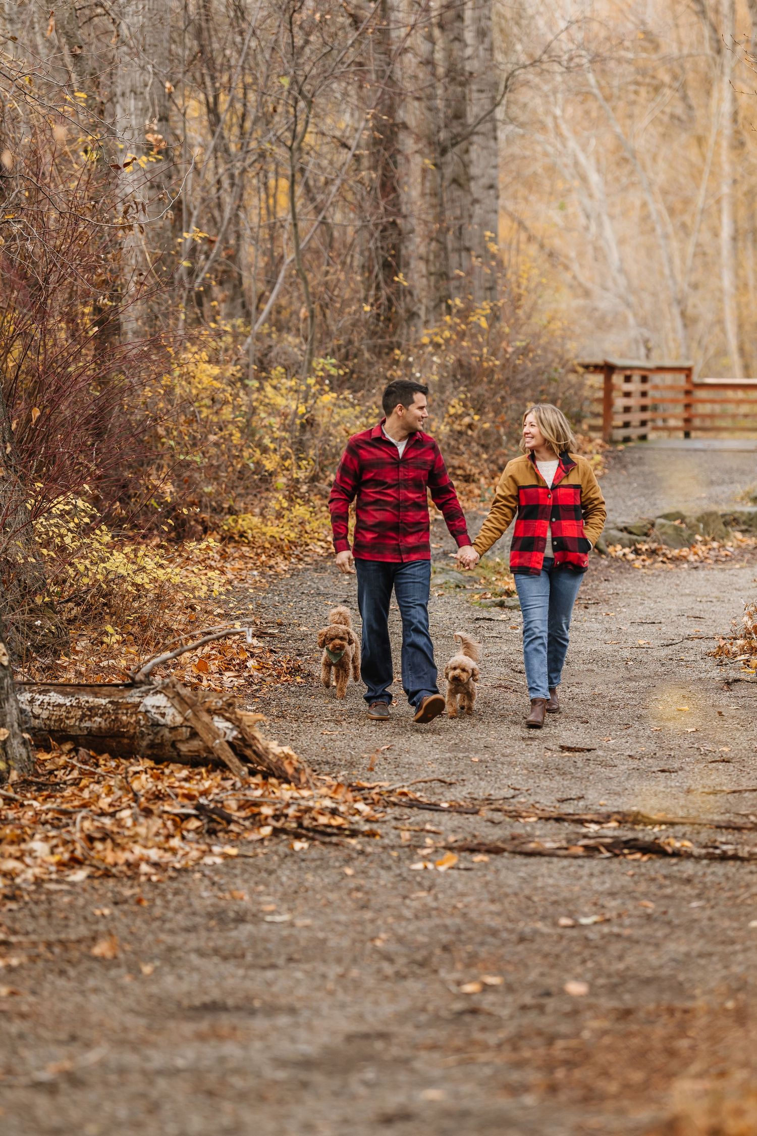 Two people in red plaid shirts stroll down a leaf-covered path in a wooded area during fall.
