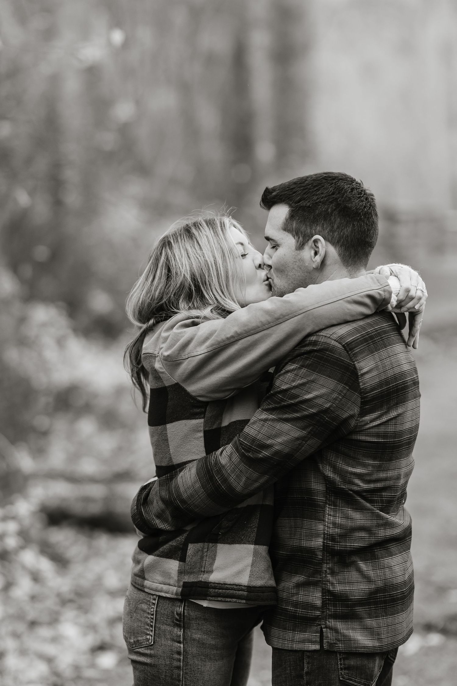 A romantic black and white photo of a couple embracing in a misty forest setting wearing plaid shirts.