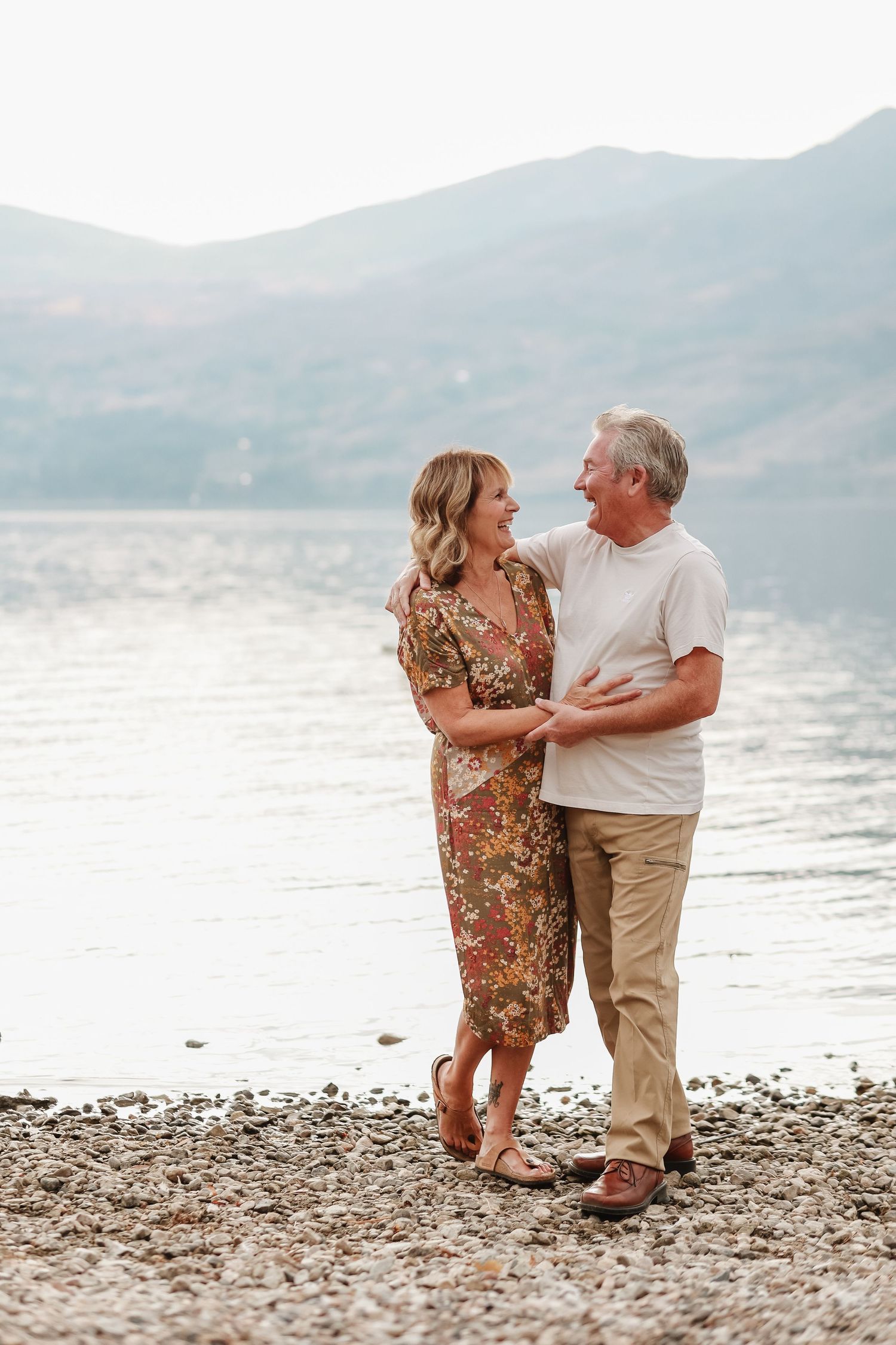 Couple dancing together on rocky shoreline with mountains and lake in soft evening light.