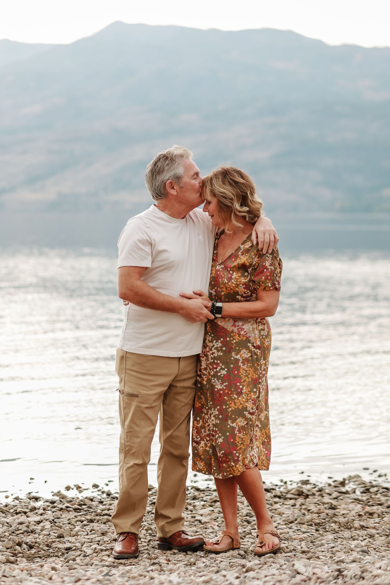Romantic couple embracing on pebble beach with mountains and lake during golden hour.