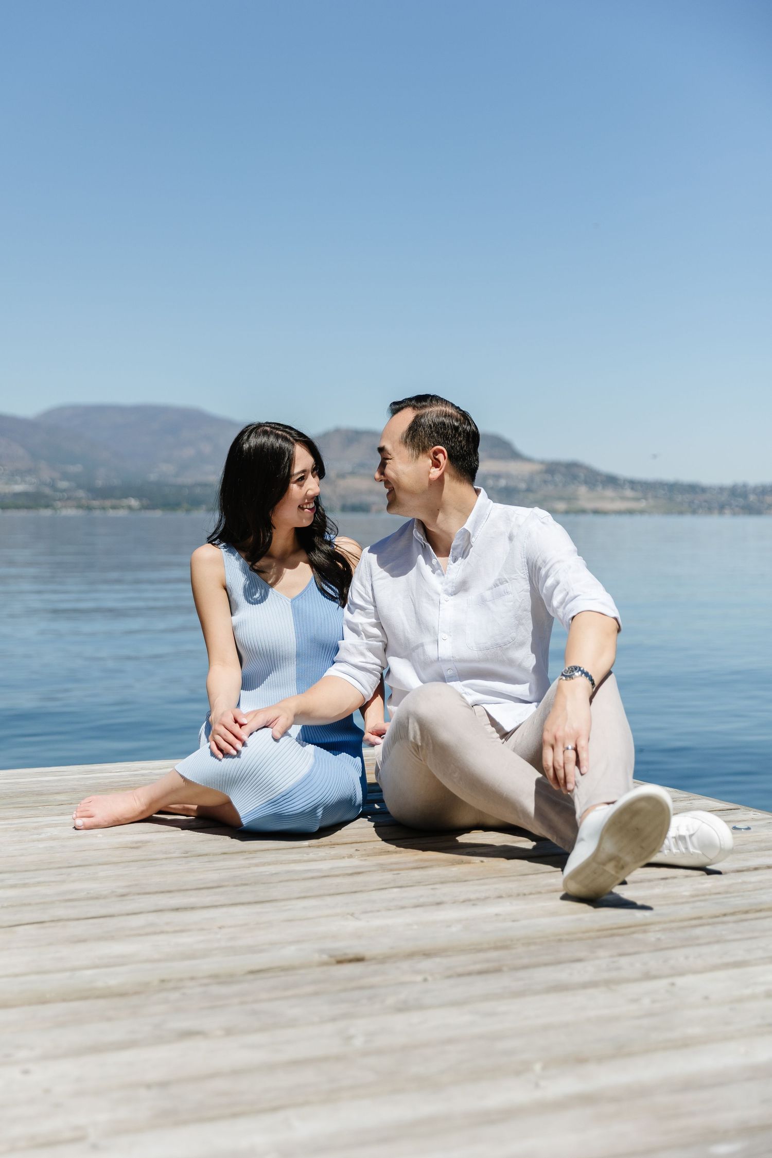 A couple relaxes together on a wooden dock overlooking a serene lake and mountain landscape.