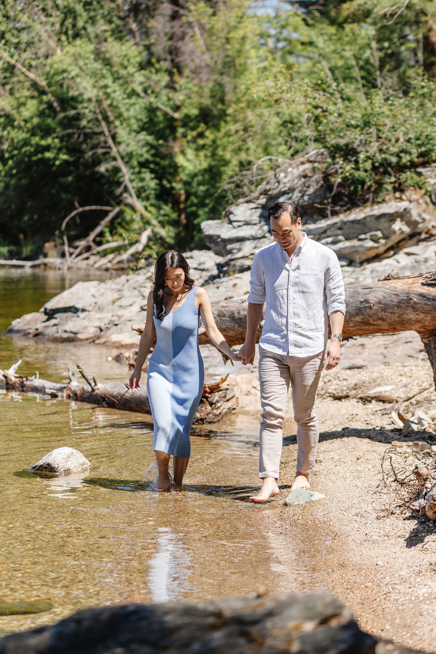 A couple in coordinated outfits takes romantic photos along a scenic riverbank with rocks and trees.