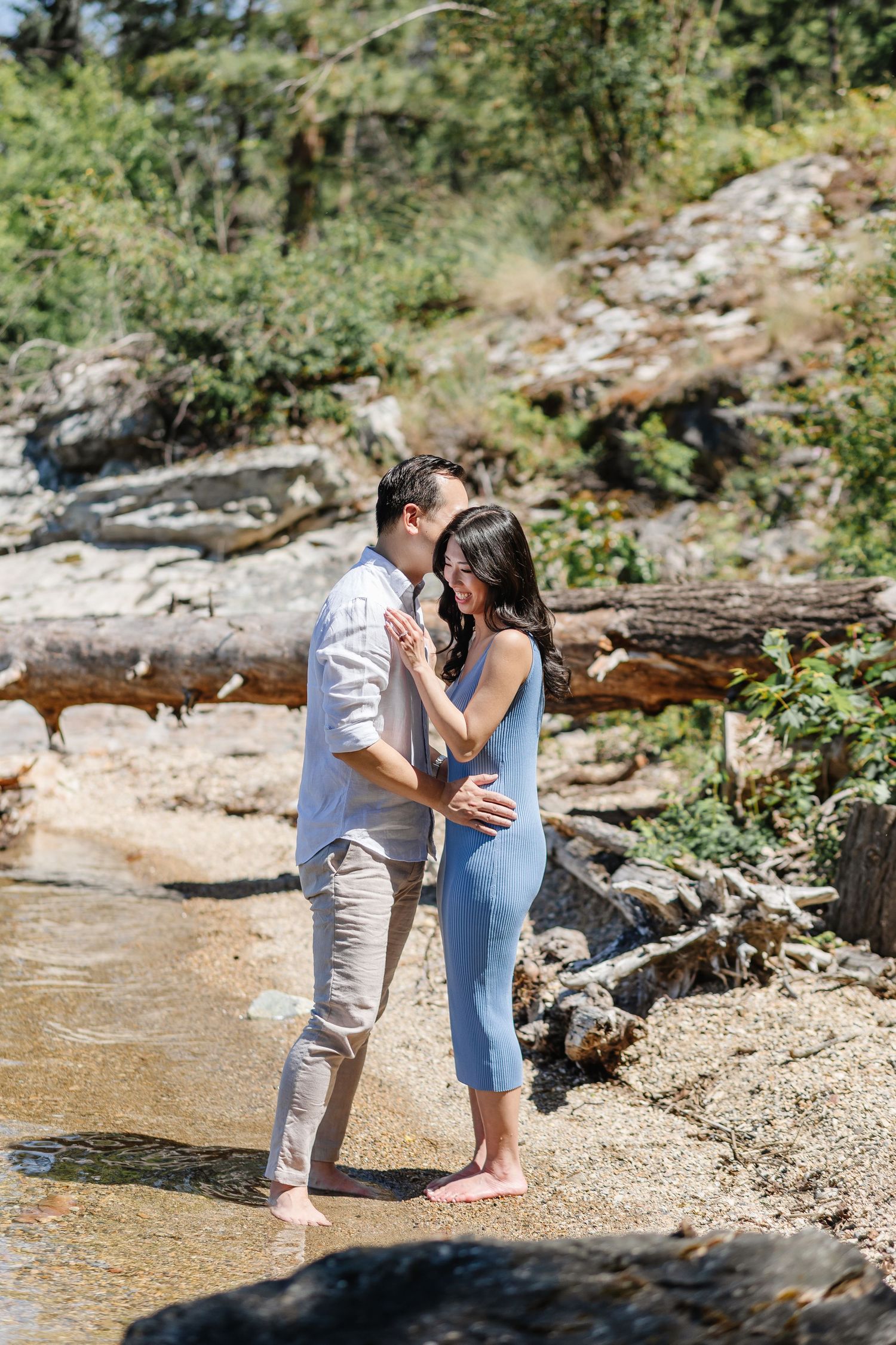 A couple shares an intimate moment by the water's edge on a rocky beach surrounded by nature.