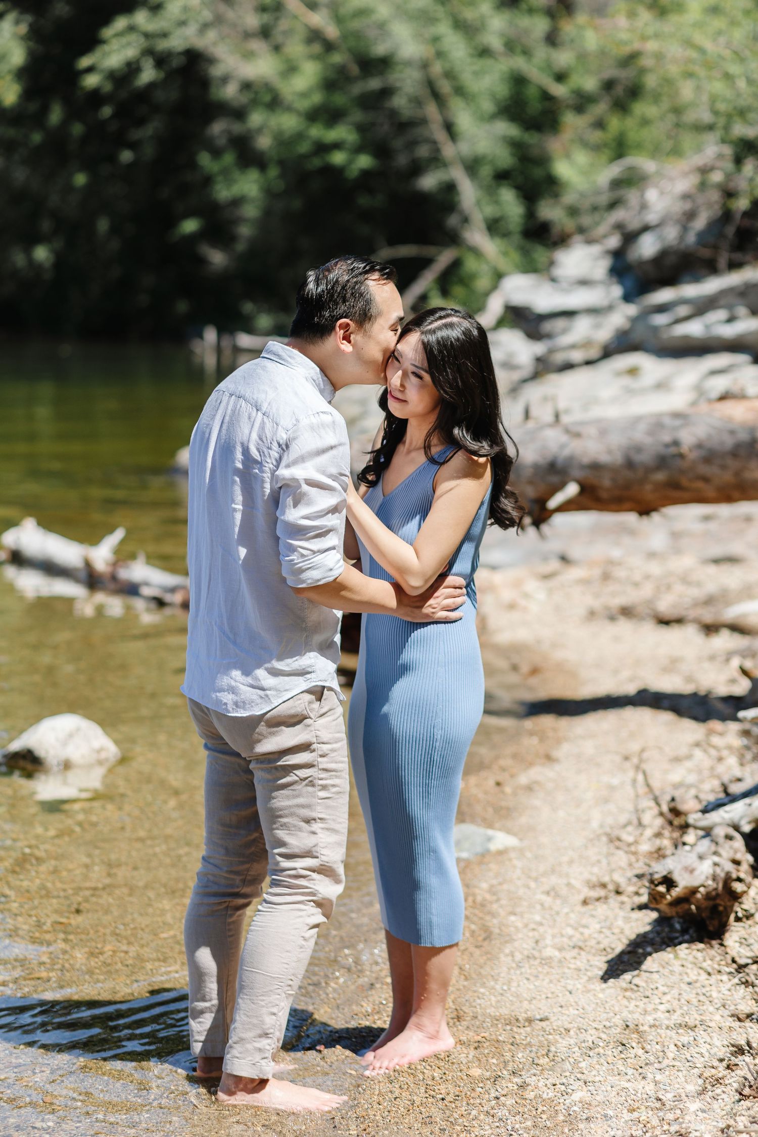 A couple embraces near the shoreline of a lake with trees and rocks in the background.