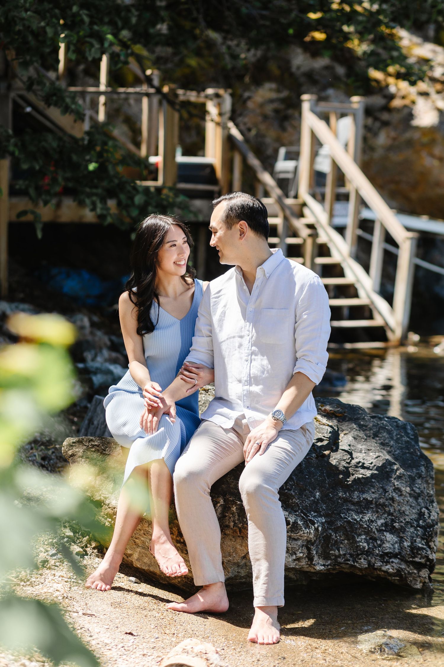 Romantic couple in white outfits sitting on rocky beach near wooden stairs in dappled sunlight.