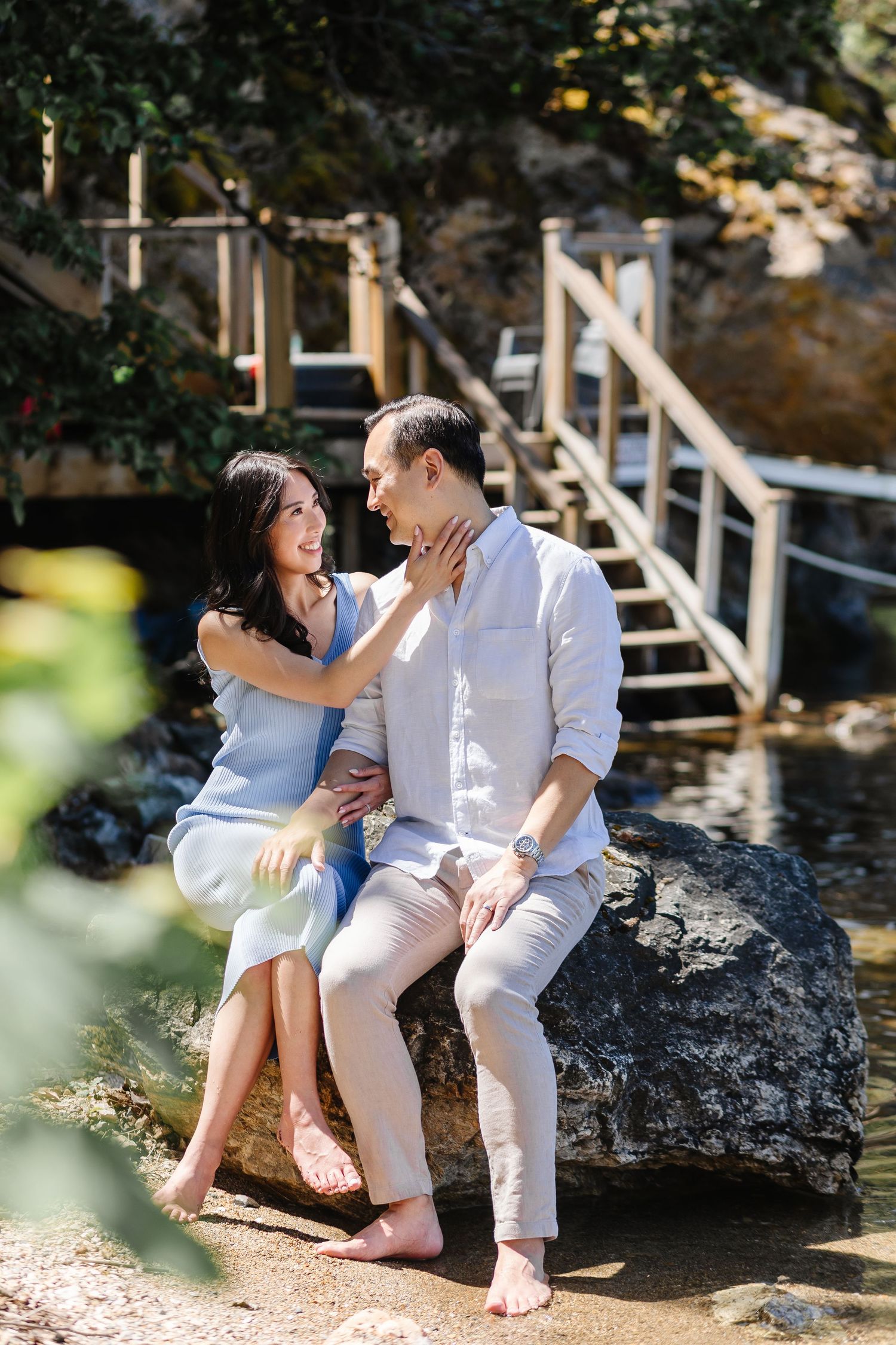 A couple shares tender moments while sitting on rocks by the water's edge in a romantic seaside photoshoot.