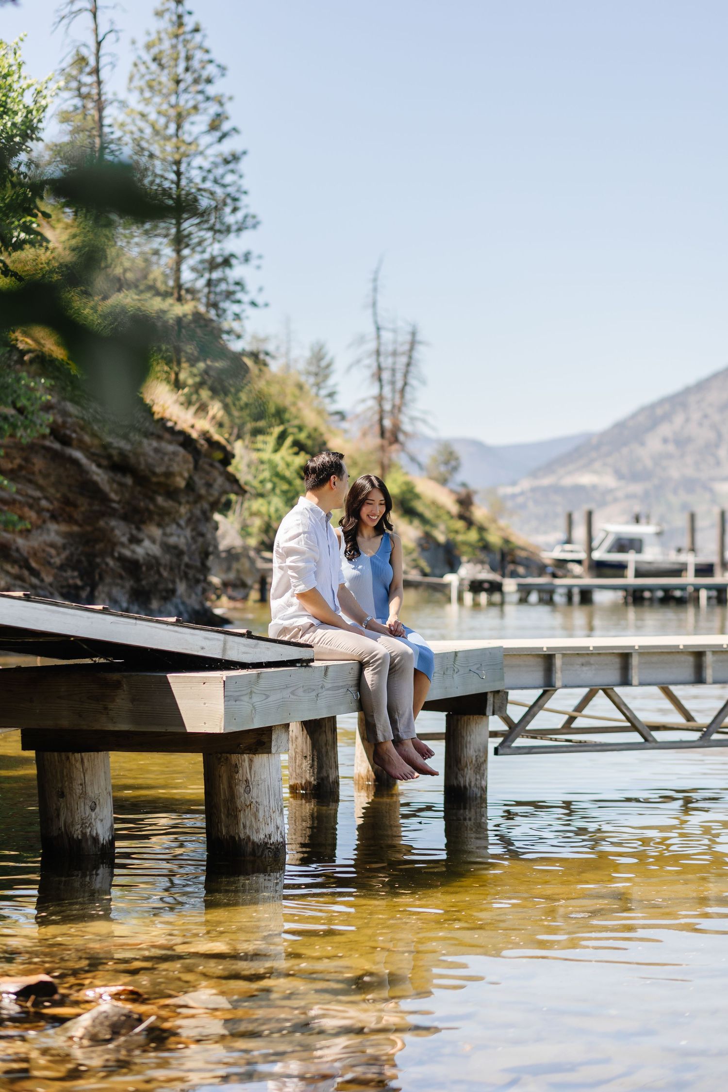 Couple sitting together on wooden dock by mountain lake with feet dangling over calm water on sunny day.