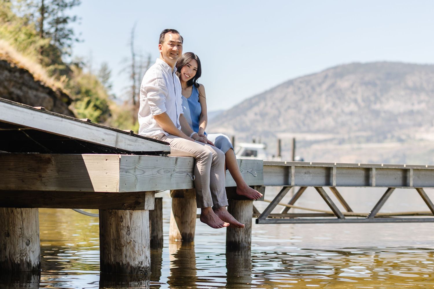 Couple sitting together on a wooden dock by a lake with mountains in the background during sunset.