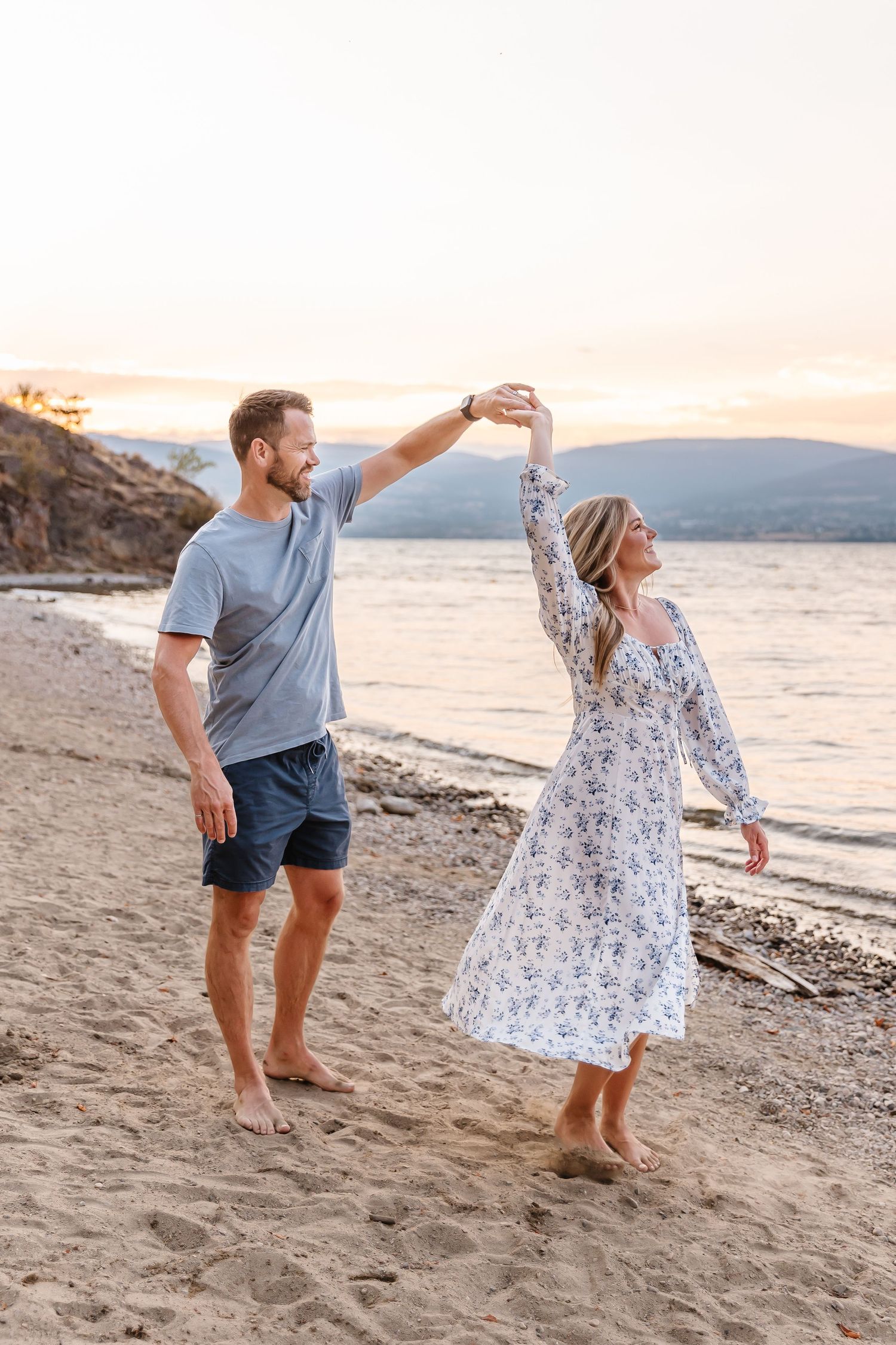 Couple dancing playfully on sandy beach at sunset with mountains in background.