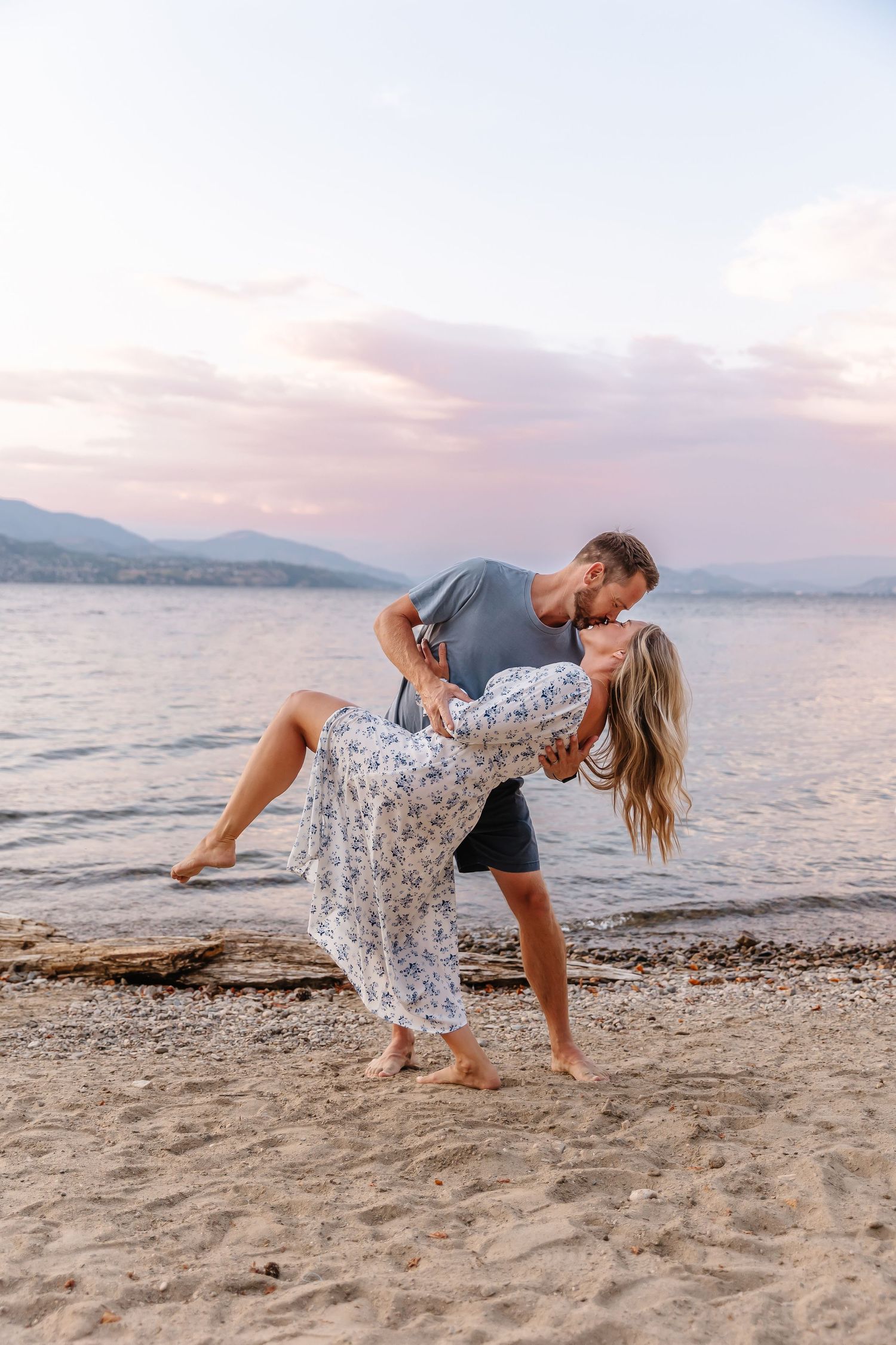 Romantic couple dances playfully on a beach at sunset with mountains in the background.