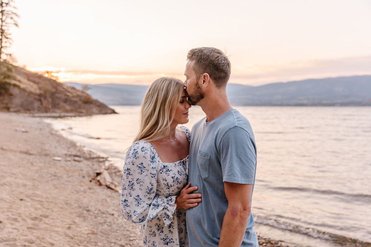 Couple embracing on sandy beach during golden hour with ocean and mountains in view.