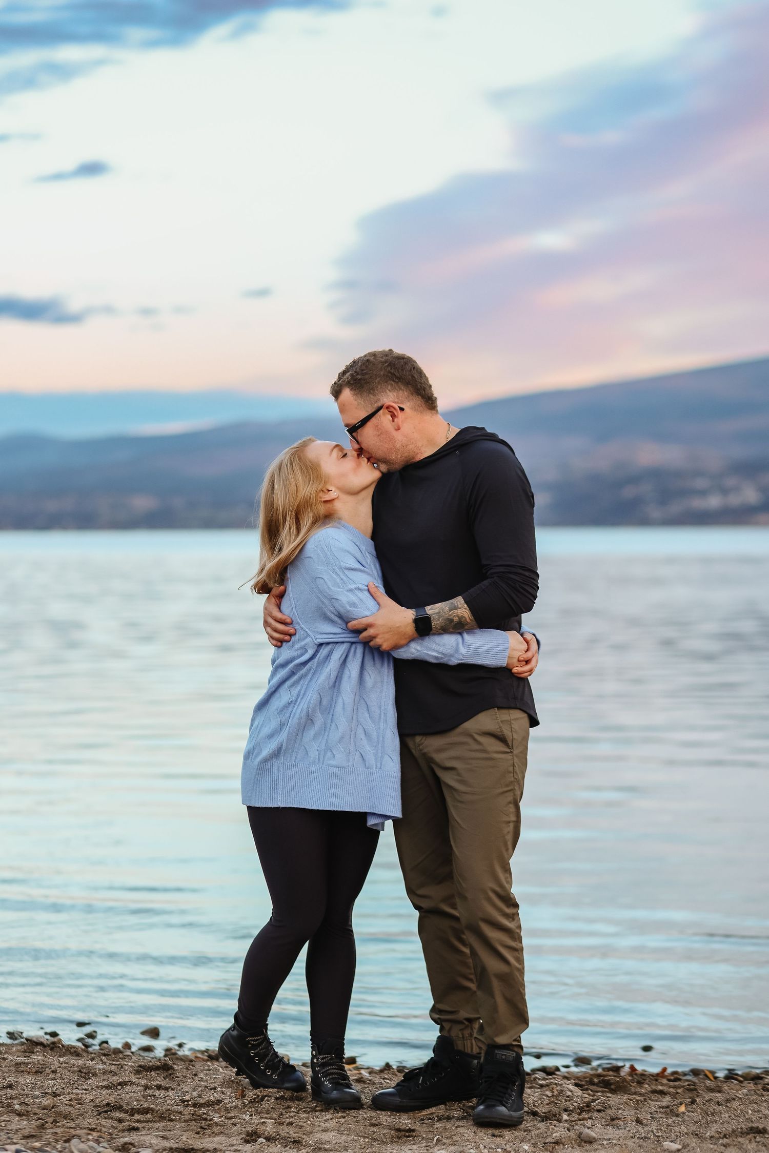 Romantic couple shares an intimate moment at sunset on a rocky beach with mountains in the background.