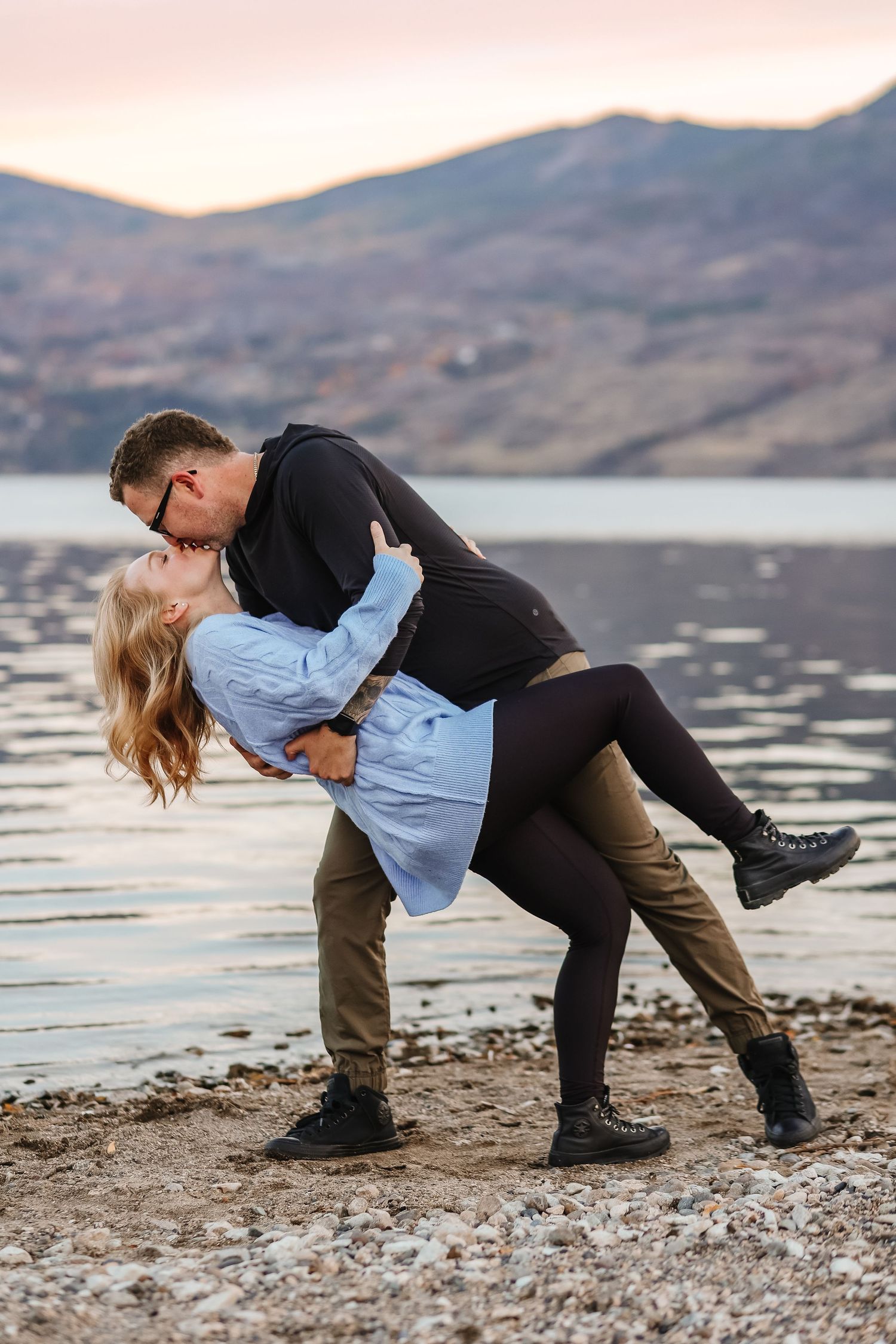 Couple performs romantic dip pose on pebble beach with mountain lake backdrop at dusk.